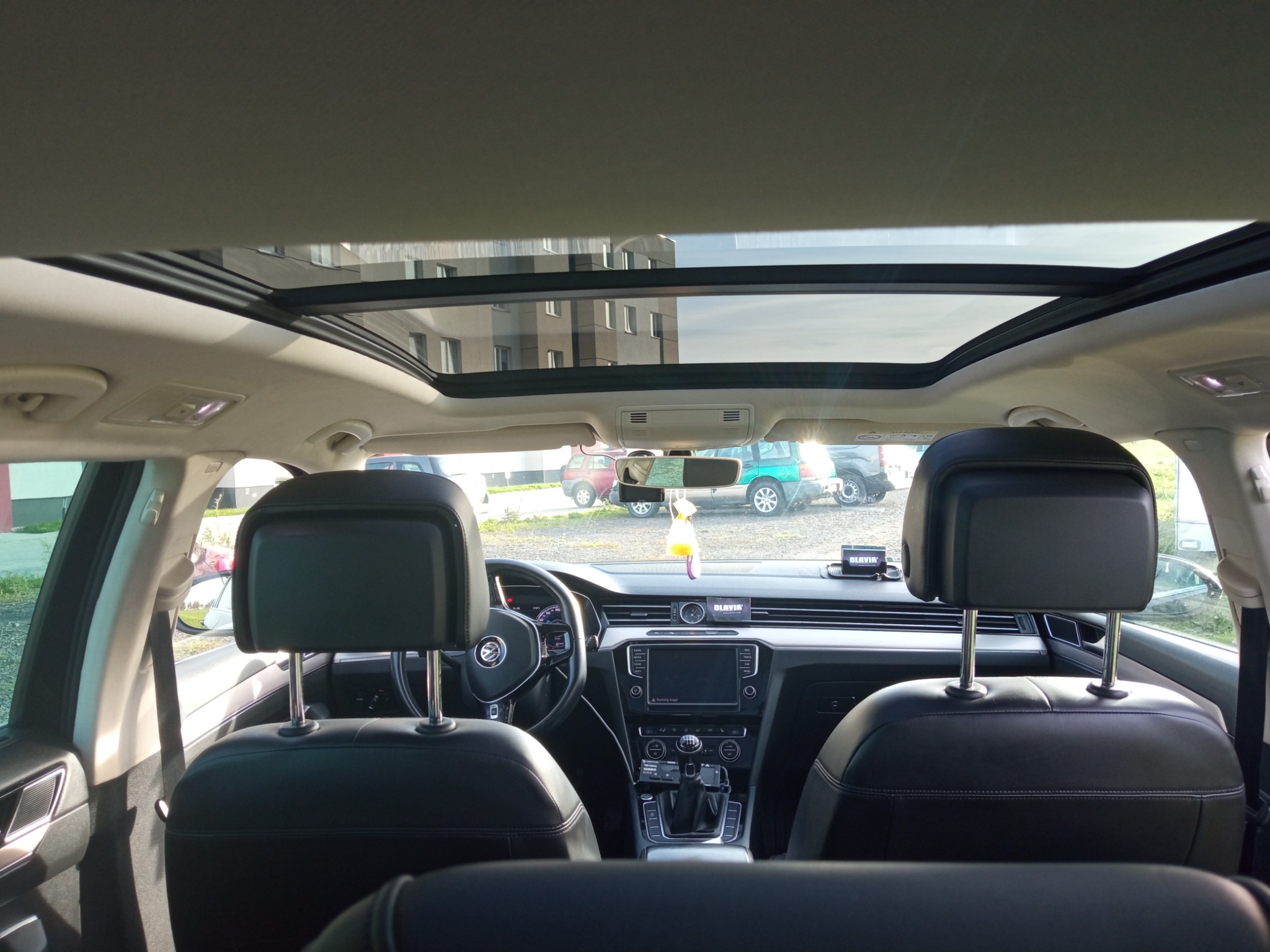 Wnętrze samochodu z widocznym panoramicznym dachem, czarną skórzaną tapicerką foteli, deską rozdzielczą z ekranem dotykowym i budynkiem w tle przez szybę.