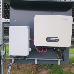 Usługi Elektryczne Witold Kornaga - Biały inwerter Sofar Solar zamontowany na konstrukcji paneli słonecznych, z widoczną skrzynką przyłączeniową i okablowaniem na tle zielonej trawy.