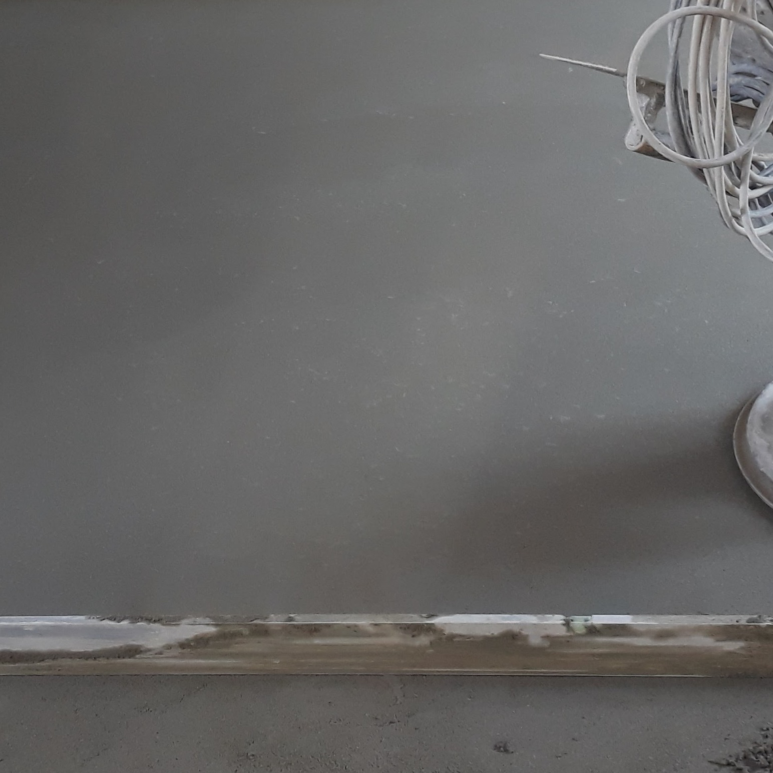 Wyrównana, szara powierzchnia posadzki jastrychowej z widoczną listwą przypodłogową i fragmentem maszyny do zacierania. Gładka, jednolita struktura.