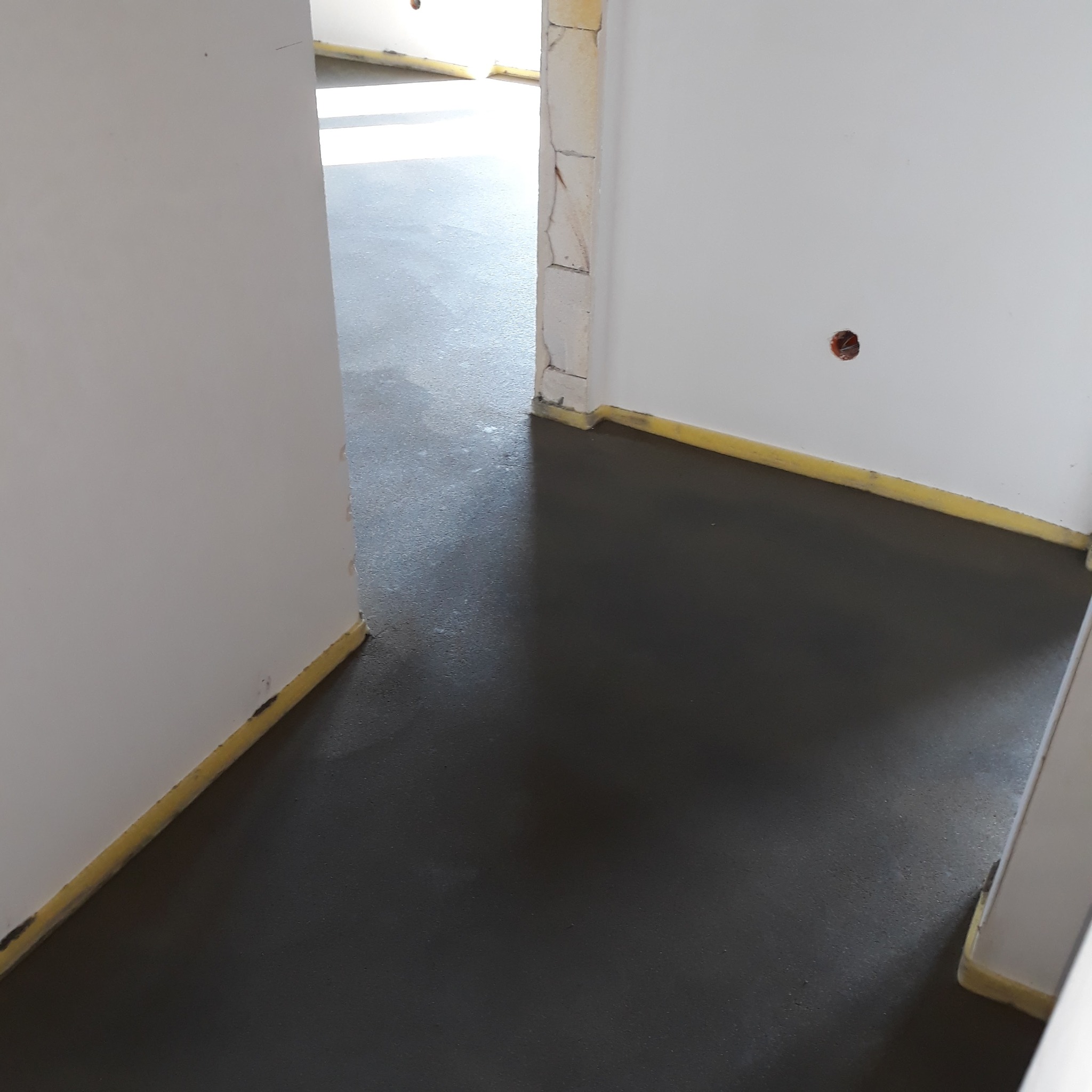 Świeżo wylana, ciemna posadzka jastrychowa w jasnym pomieszczeniu z białymi ścianami. Żółta taśma zabezpiecza ściany. Widok z góry na gładką powierzchnię.
