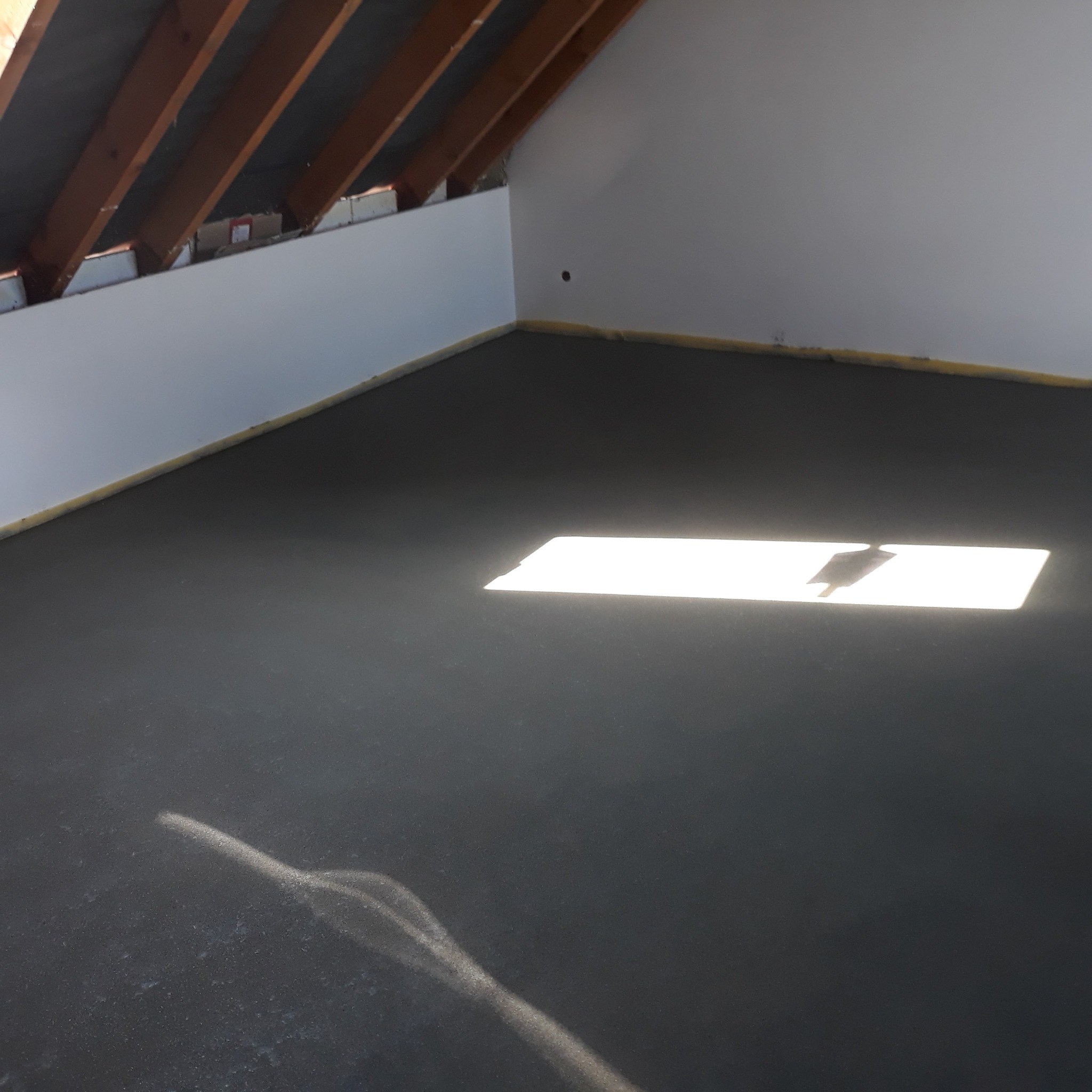 Świeżo wylana, ciemna posadzka jastrychowa w jasnym pomieszczeniu poddasza z drewnianą konstrukcją dachu. Na powierzchni widoczny prostokątny refleks światła.