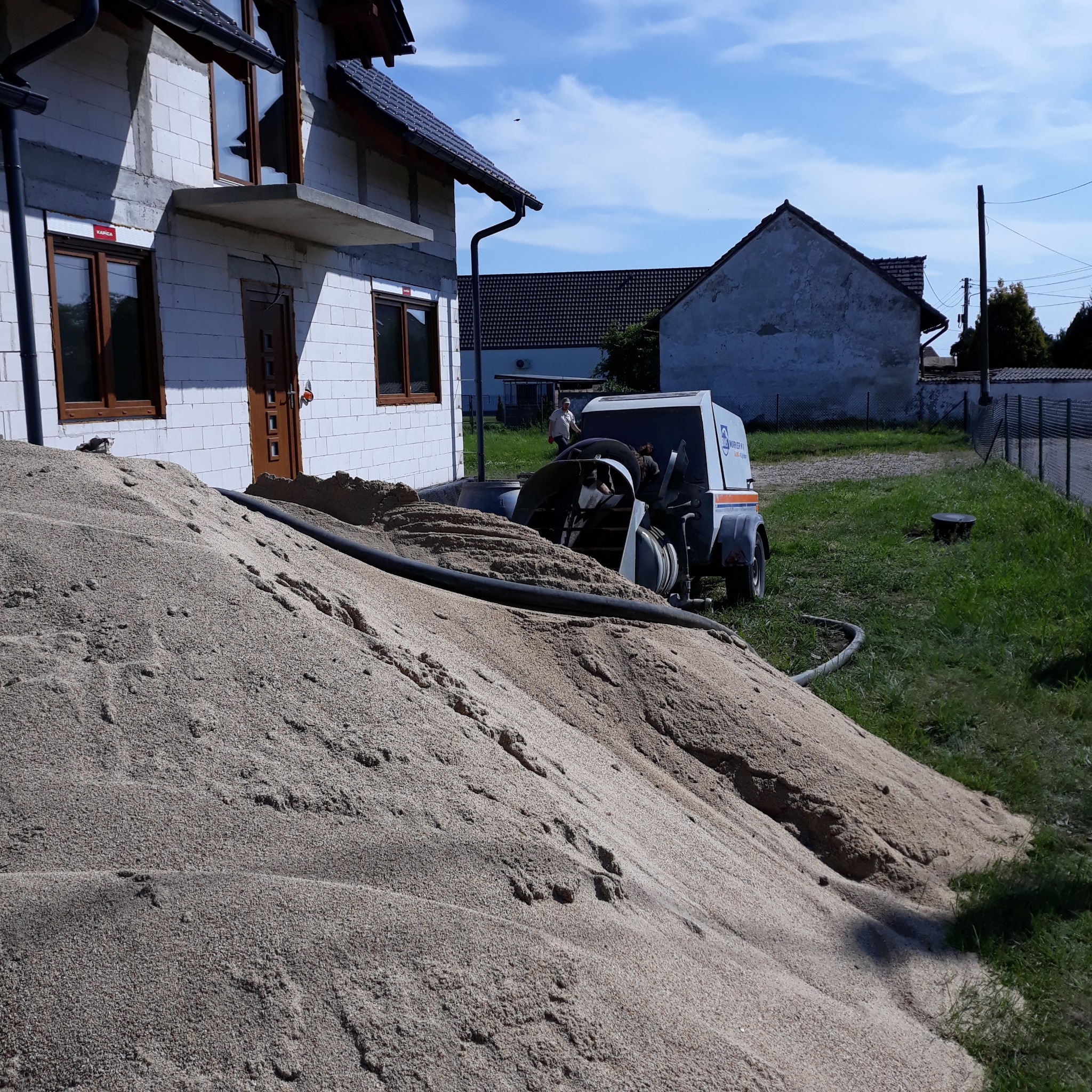 Stos piasku obok domu w trakcie budowy, z maszyną do tynkowania w tle. Widoczny fragment elewacji z białego bloczka i okna z brązowymi ramami.