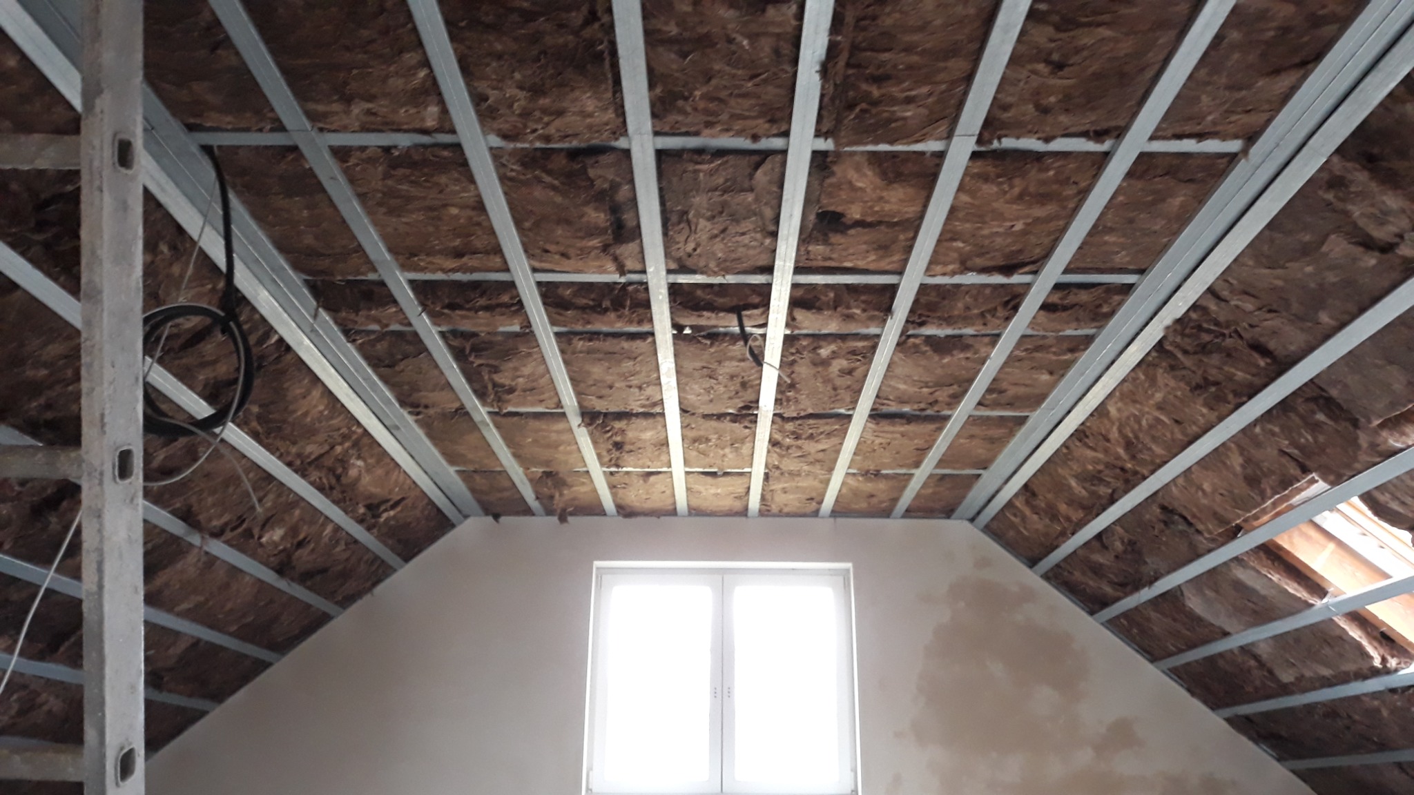 Montaż izolacji z wełny mineralnej na poddaszu, widoczne metalowe profile konstrukcyjne. Okno dachowe w tle. Prace wykończeniowe w trakcie realizacji.