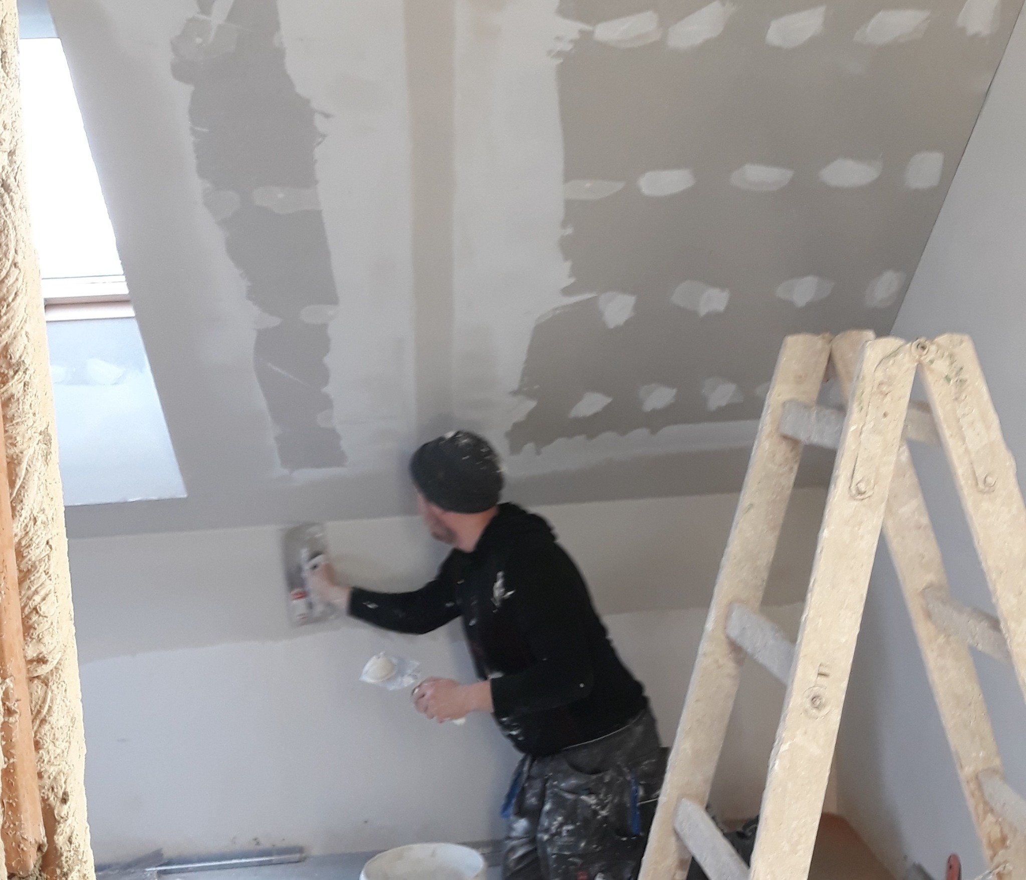 Pracownik w czarnej bluzie szpachluje ścianę na poddaszu, używając szpachelki. Widoczna drabina, okno dachowe i ślady po łatach na suficie.