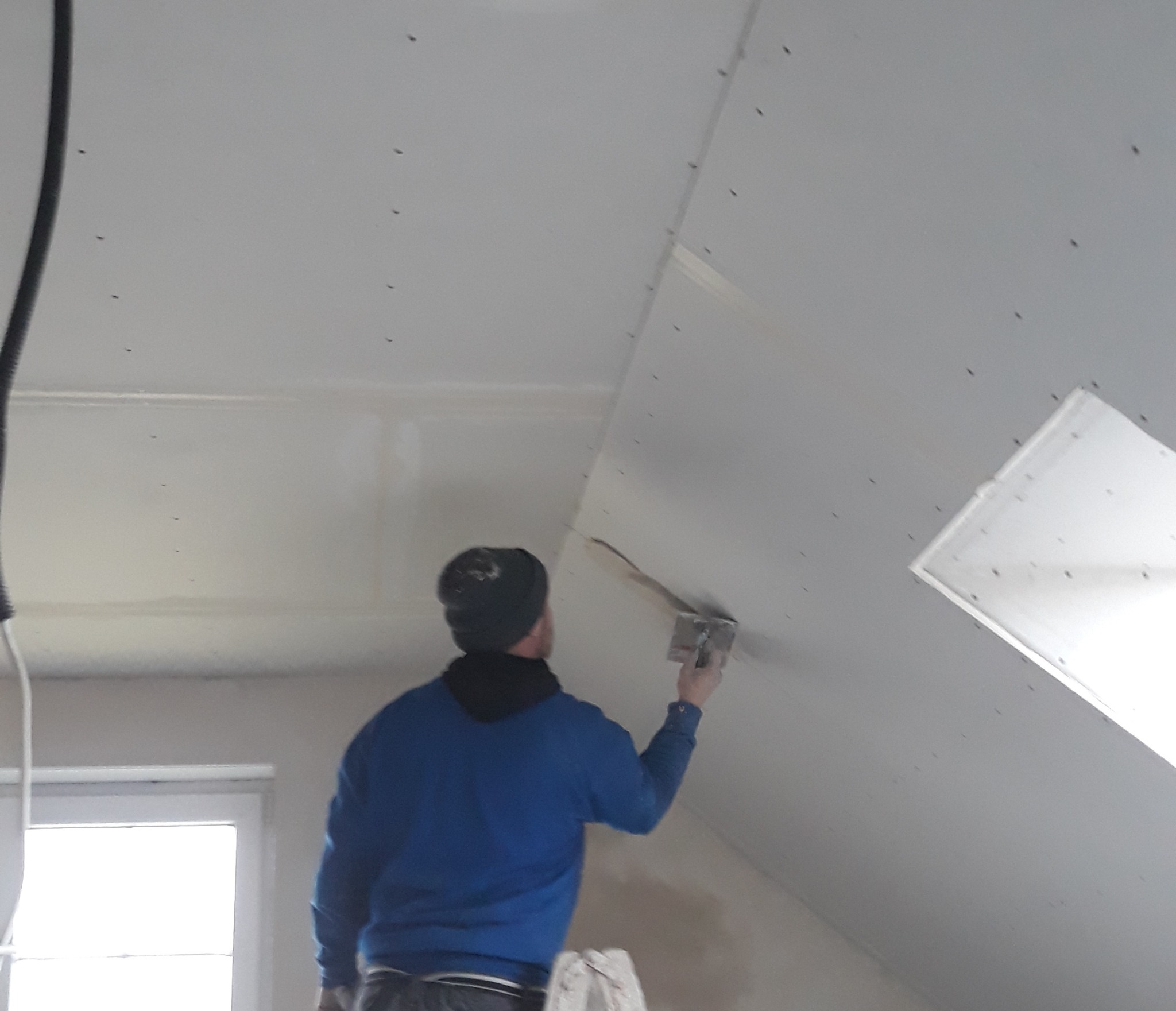 Pracownik w niebieskiej bluzie szpachluje łączenie płyt karton-gips na skośnym poddaszu z oknem dachowym. Widoczne ślady po wkrętach.