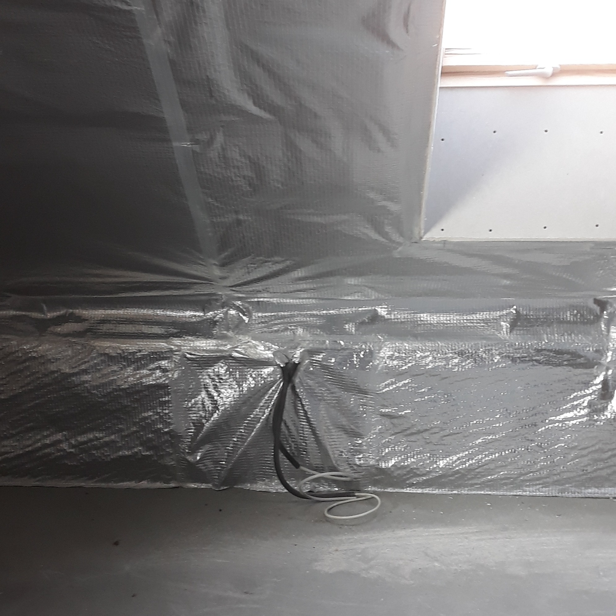 Izolacja termiczna poddasza z folią aluminiową, widoczne przewody elektryczne. Ściana z płyty gipsowej w tle. Ujęcie z dołu, podkreślające detale izolacji.