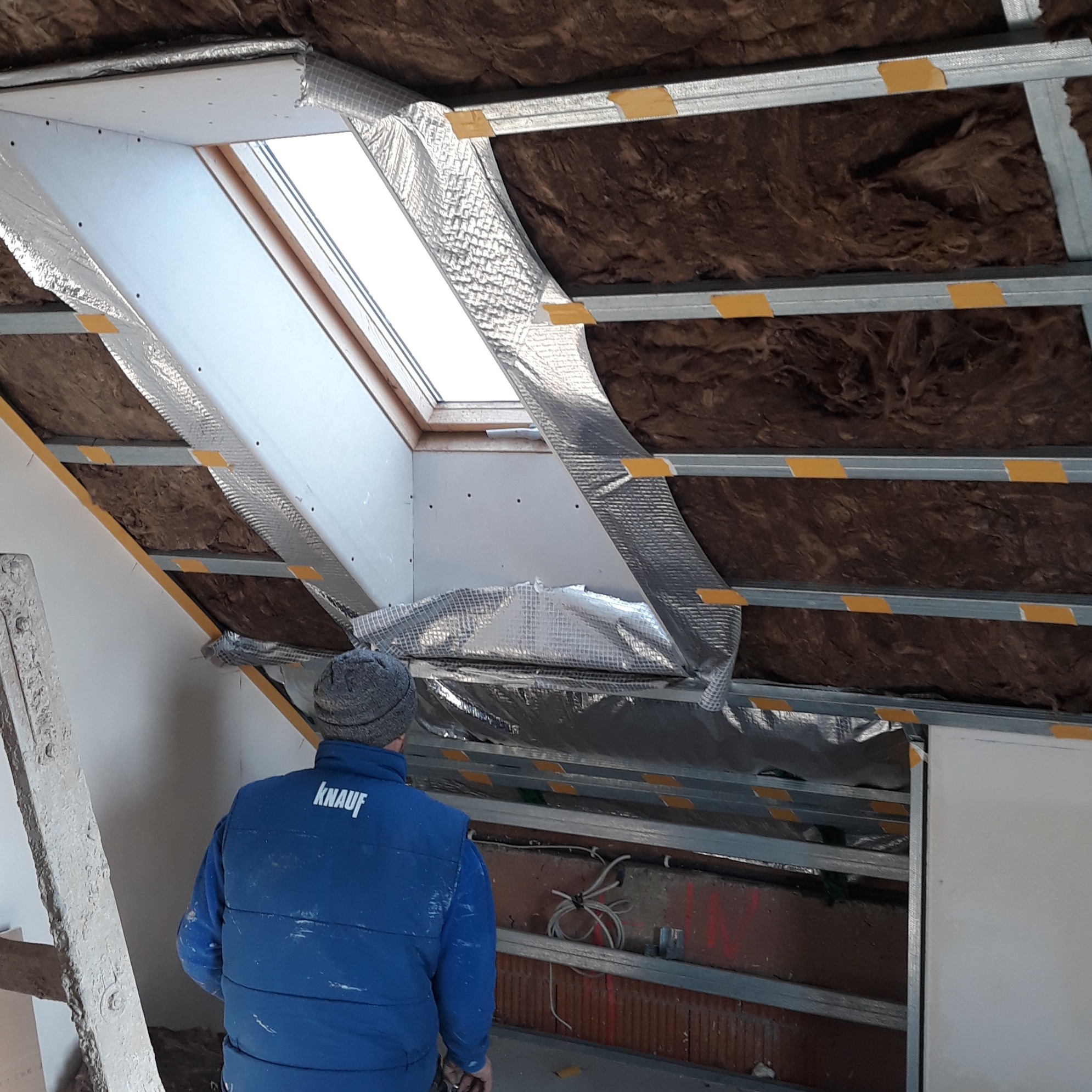 Montaż izolacji poddasza z oknem dachowym w Otmicach. Widoczna wełna mineralna, folia aluminiowa i konstrukcja z profili. Pracownik w ubraniu roboczym firmy Knauf.
