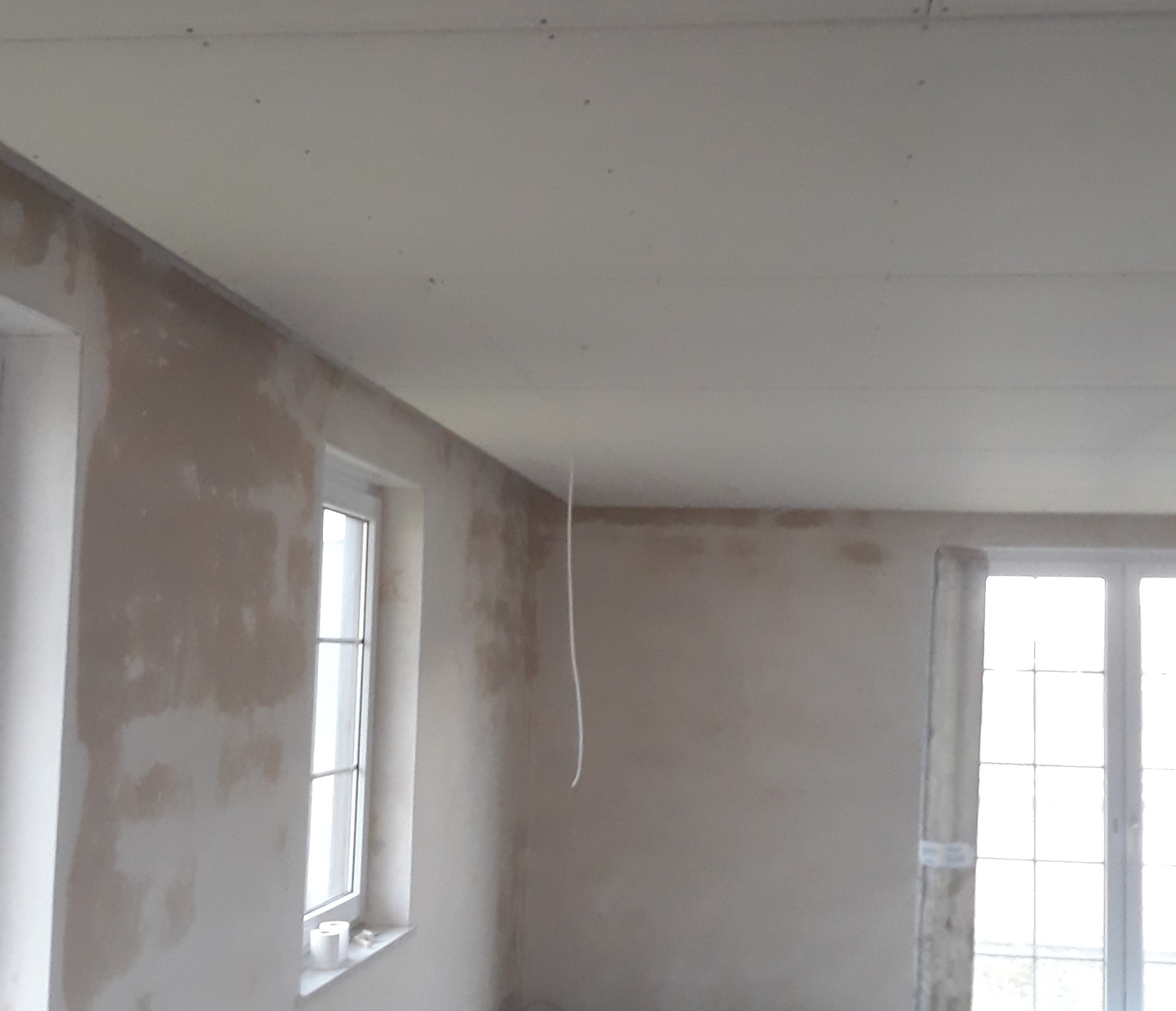 Pokój w trakcie prac wykończeniowych: ściany z widocznymi śladami szpachlowania, sufit z płyt kartonowo-gipsowych, okna z białymi ramami. W tle widoczne kolejne okno.