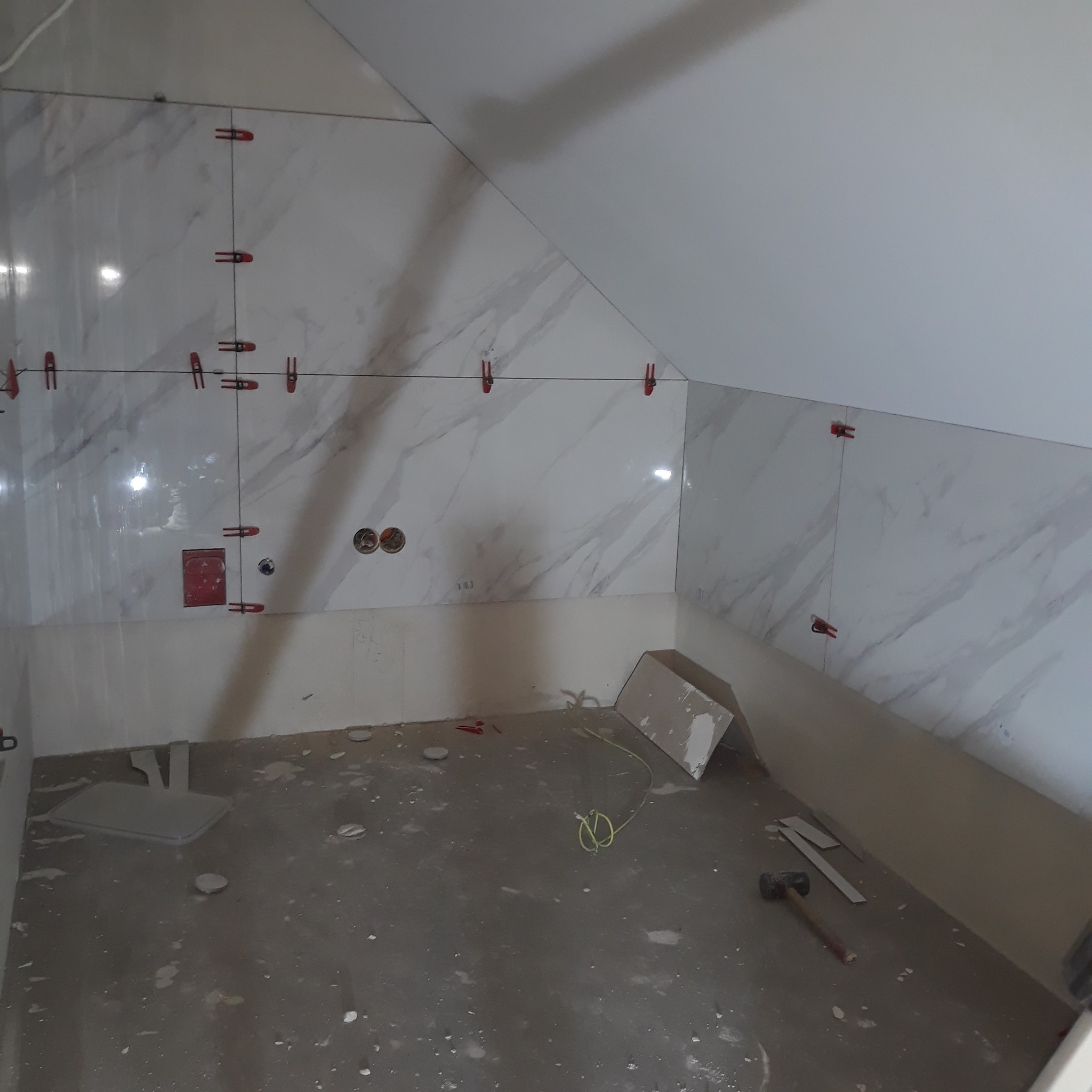 Ściany w trakcie układania płytek imitujących marmur, z użyciem klipsów poziomujących. Widoczne otwory na gniazdka i fragment podłogi z narzędziami.