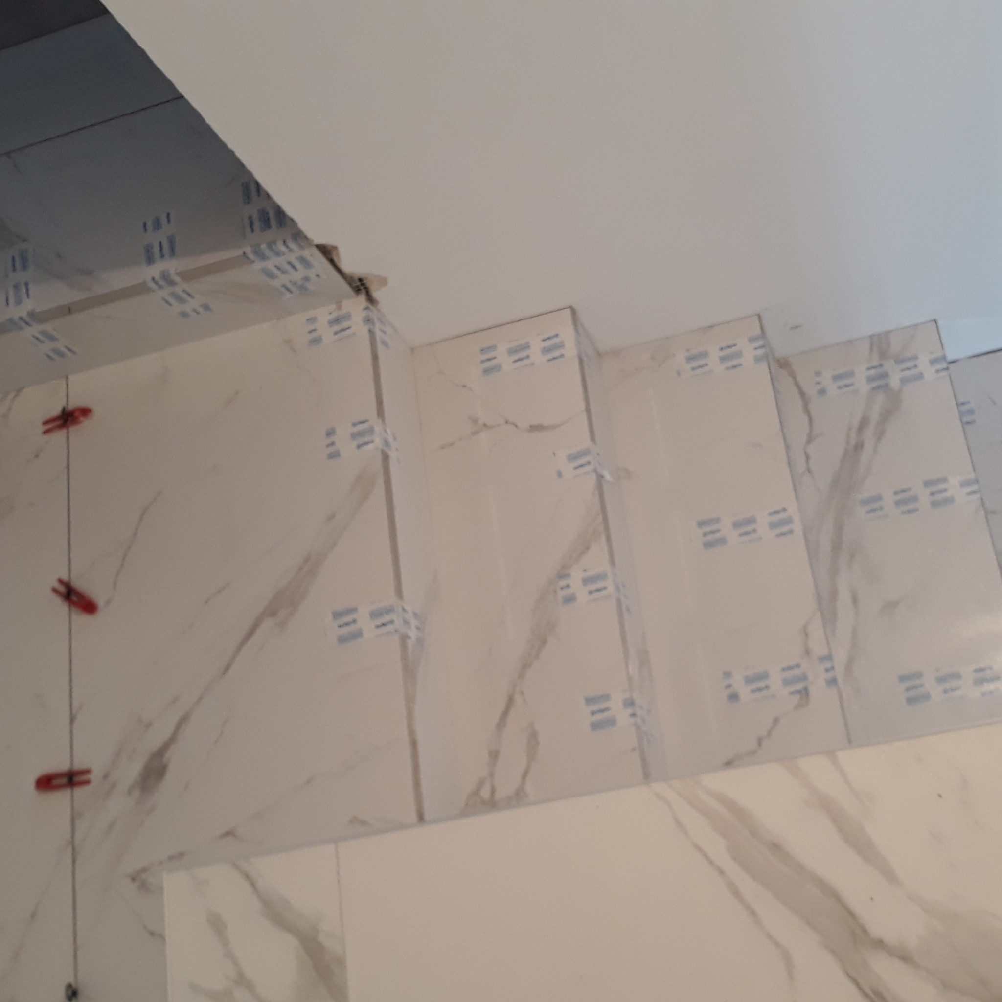 Układanie płytek imitujących marmur na ścianie, widoczne krzyżyki dystansowe i taśmy zabezpieczające. Praca w toku, jasne wnętrze.