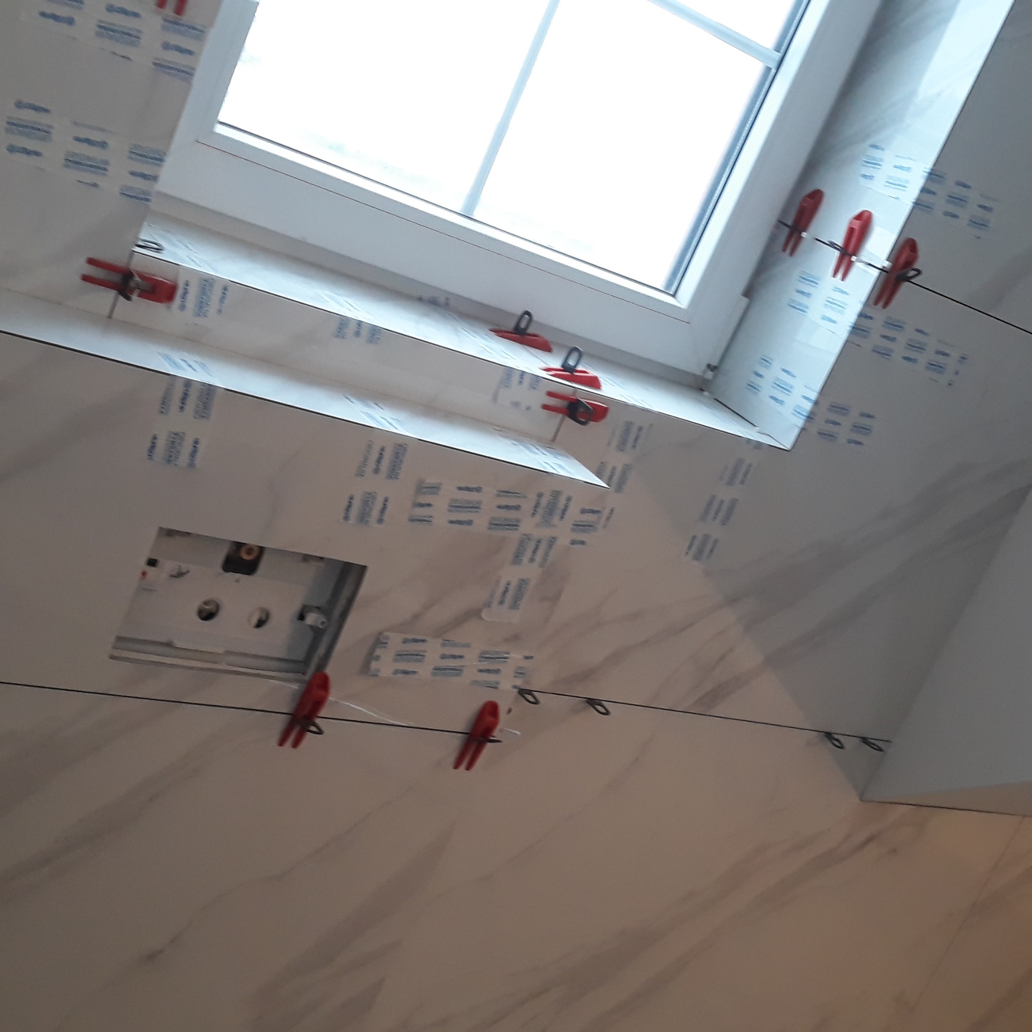 Układanie płytek imitujących marmur w łazience przy oknie dachowym, z użyciem klipsów poziomujących. Widoczny otwór na instalację. Praca w toku.