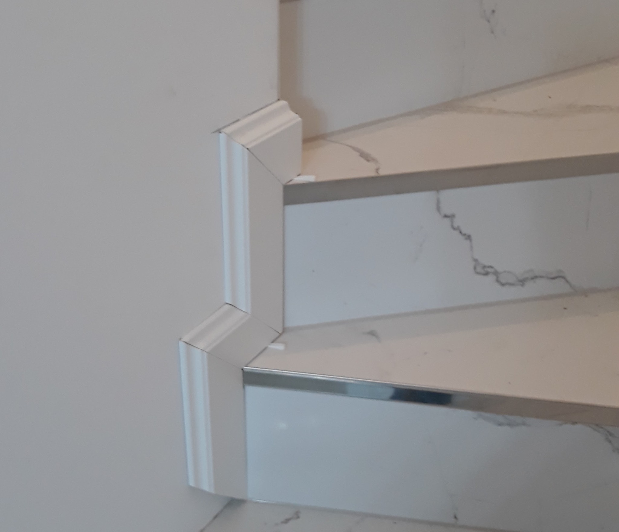 Wykończenie schodów z marmurowymi stopniami i białą listwą przypodłogową z ozdobnym frezem. Detal wykończeniowy z metalową krawędzią. Minimalistyczny design.