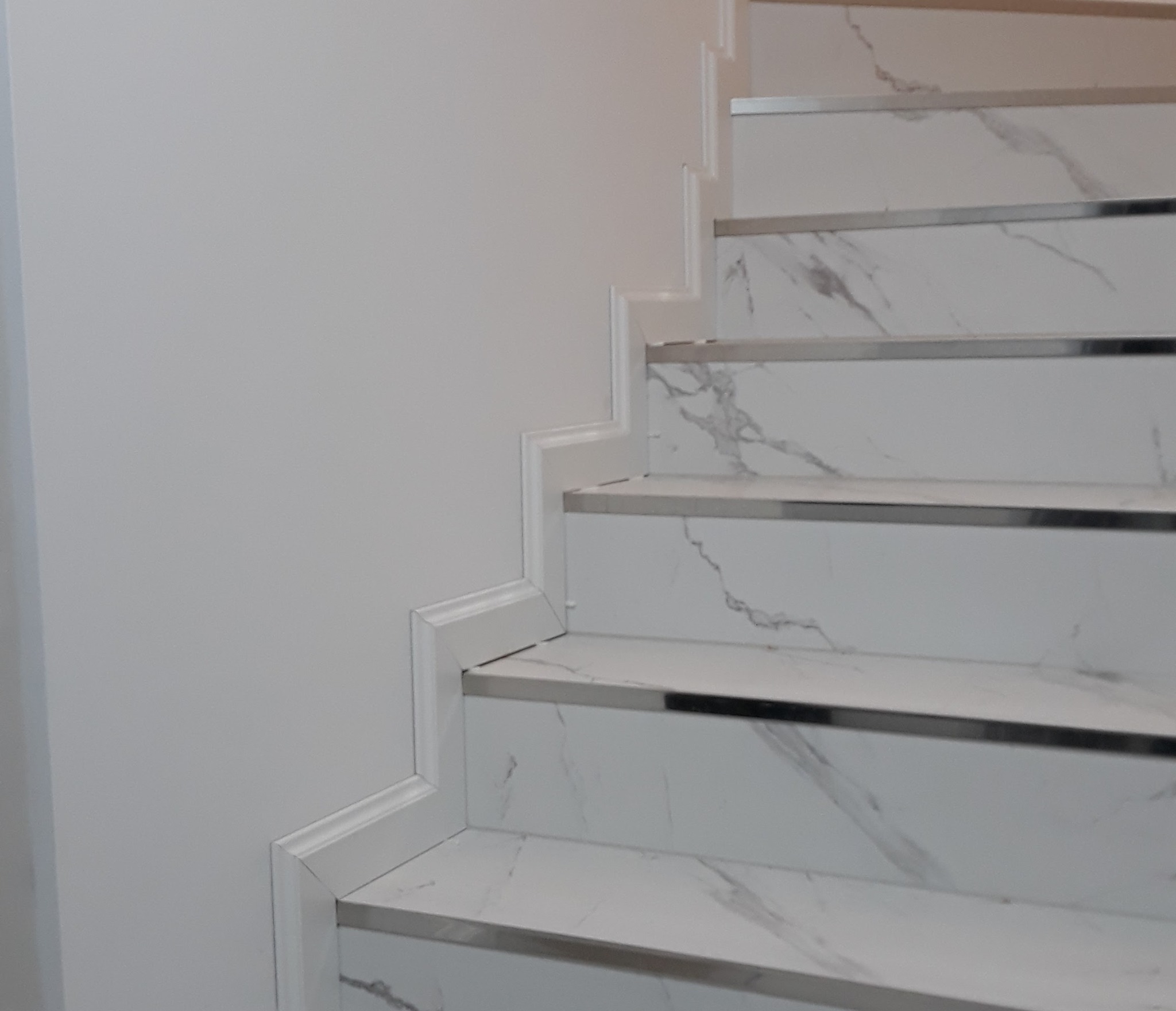 Białe marmurowe schody z metalowymi listwami, widok z dołu, ukazujące precyzyjne wykończenie i elegancki design. Detal wykończenia ściany przy schodach.