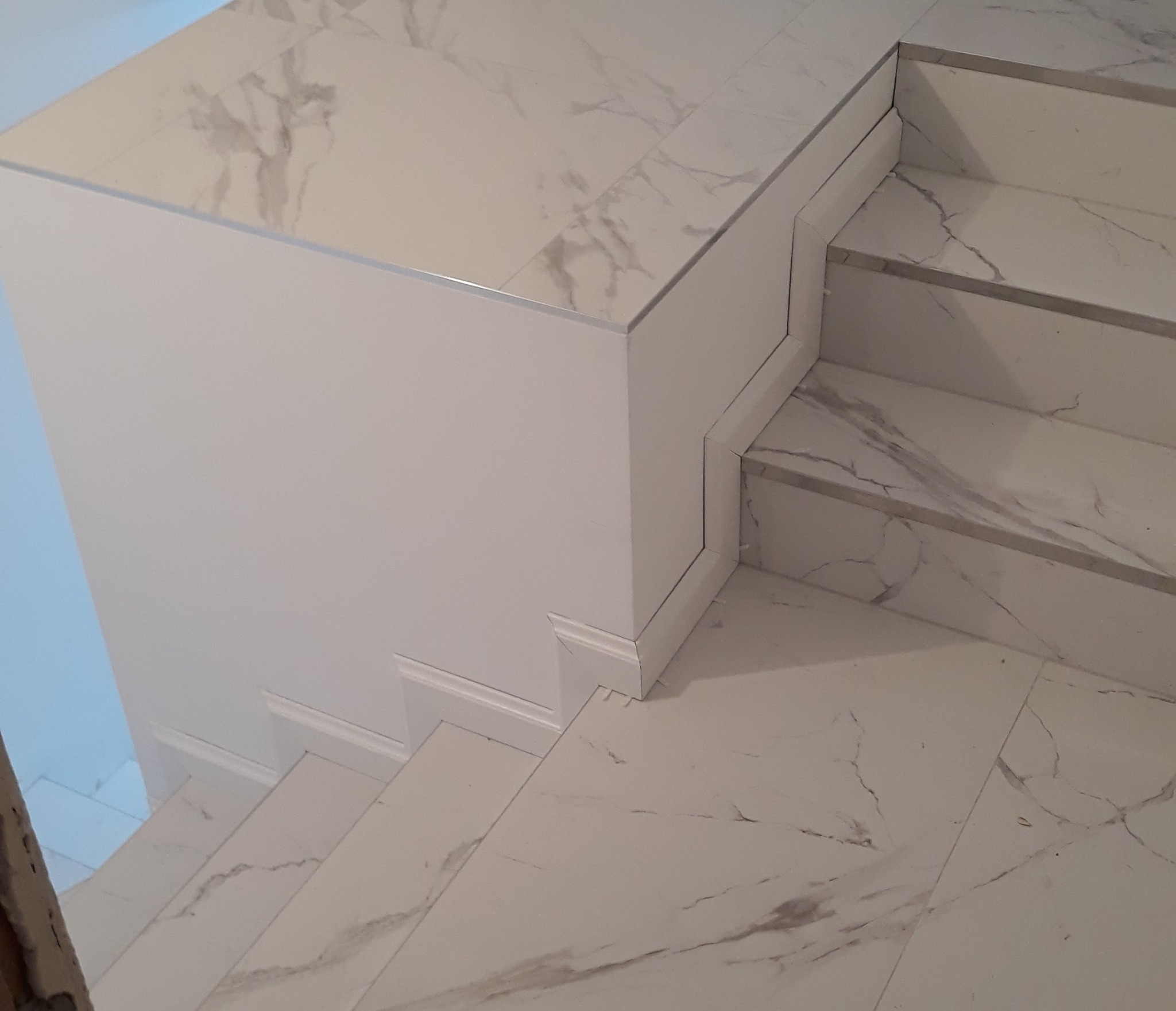 Wykończone schody z białych płytek imitujących marmur, z widocznymi detalami wykończeniowymi i listwami. Ujęcie z góry, podkreślające precyzję ułożenia.