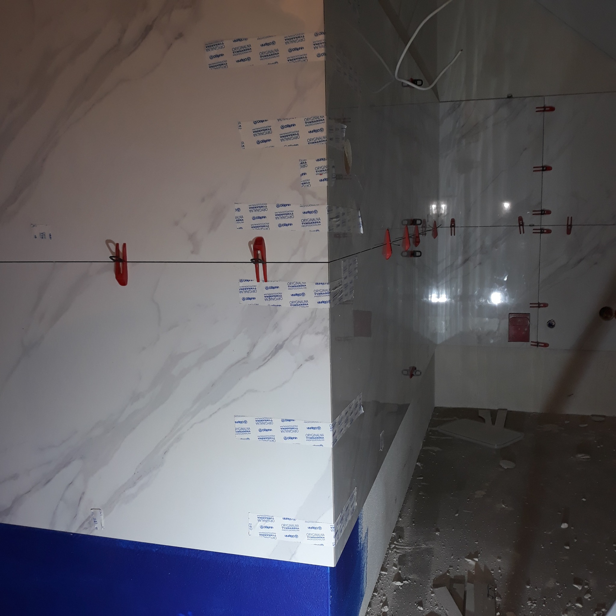 Białe płytki imitujące marmur, ułożone na ścianie z użyciem klipsów poziomujących. Widoczne taśmy zabezpieczające krawędzie. Prace glazurnicze w trakcie realizacji.