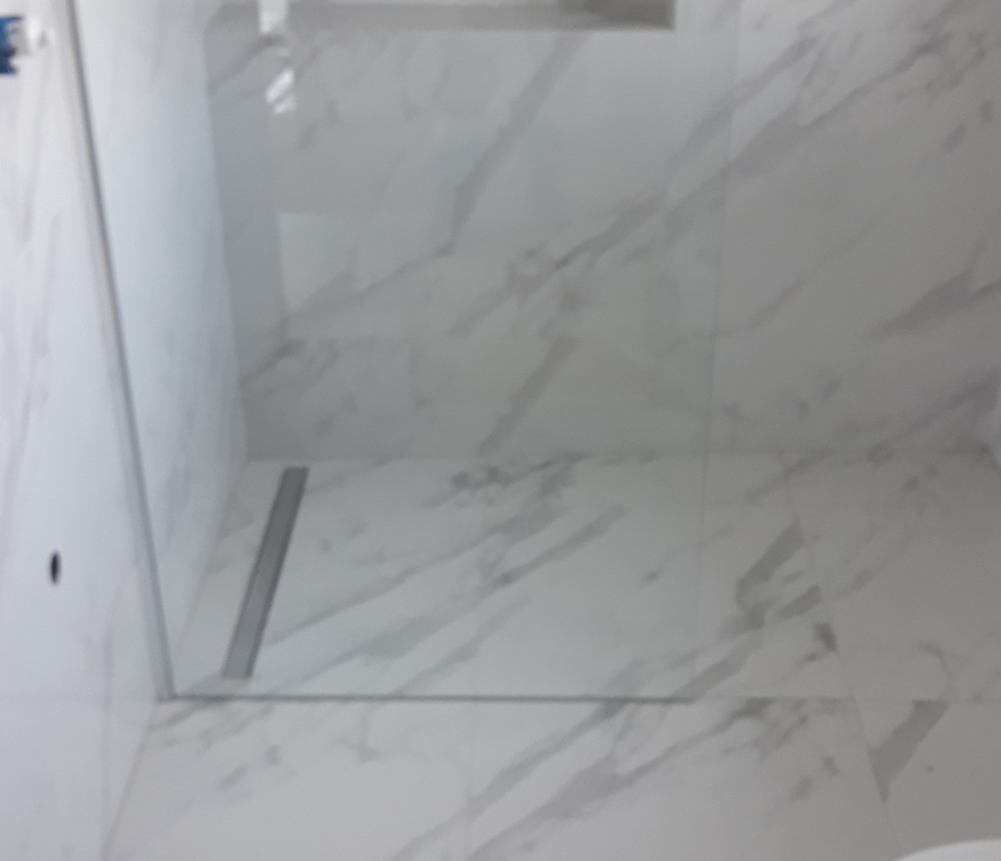 Biała łazienka wykończona płytkami imitującymi marmur, z widocznym odpływem liniowym. Minimalistyczny design, jasne wnętrze.