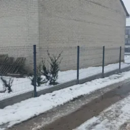 Szary panelowy system ogrodzeniowy z podmurówką na tle budynku z białej cegły i zasp śnieżnych.