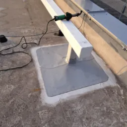 Prace dekarskie na płaskim dachu: zgrzewanie papy bitumicznej wokół podstawy konstrukcji wsporczej z użyciem zgrzewarki elektrycznej, widoczne ślady wcześniejszych prac na powierzchni dachu.