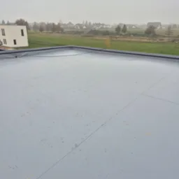 Szare pokrycie płaskiego dachu z widocznymi łączeniami, na tle zabudowań i zamglonego krajobrazu.