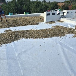 Pokrycia Dachowe Adrian Szczyrba - Biała membrana dachowa z rozłożonym żwirem na dachu w Zabrzu, widoczne narzędzia i materiały budowlane, drzewa w tle.