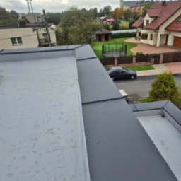 Szary, płaski dach z widocznymi obr&oacute;bkami blacharskimi w kolorze grafitowym. W tle zabudowa jednorodzinna i kości&oacute;ł. Widok z g&oacute;ry na nową konstrukcję dachu.