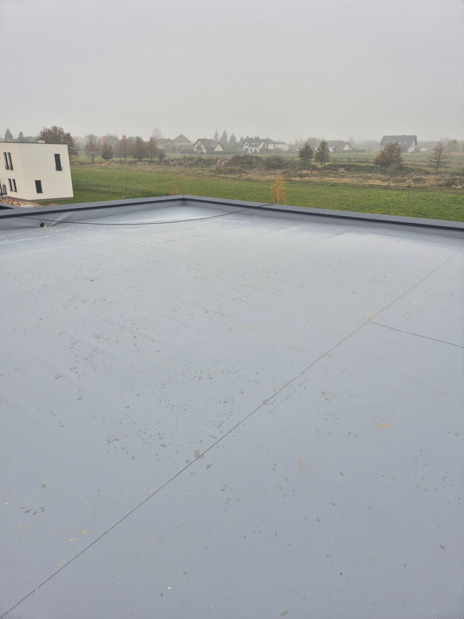 Szare pokrycie płaskiego dachu z widocznymi łączeniami, na tle zabudowań i zamglonego krajobrazu.