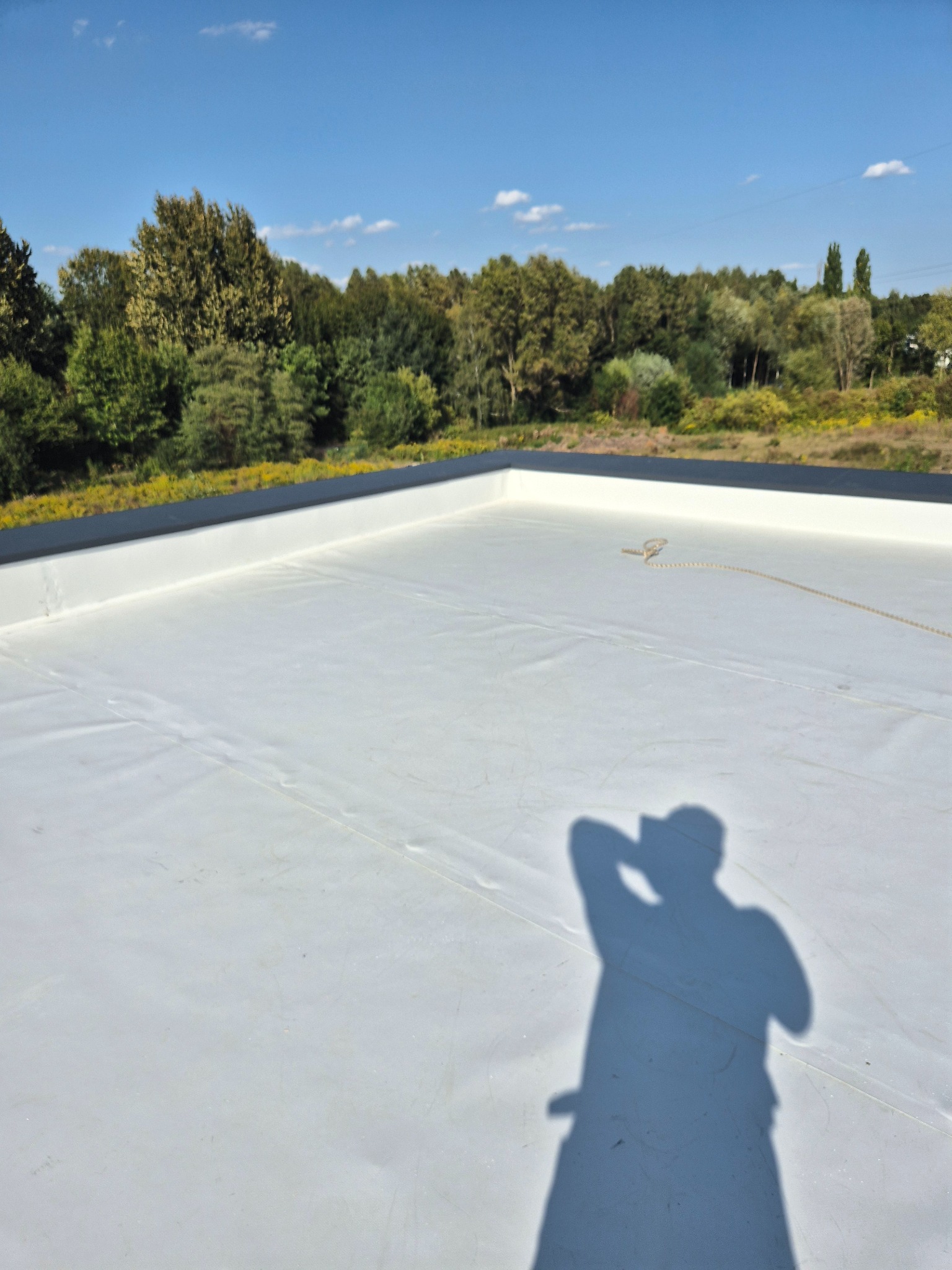 Biała, płaska powierzchnia dachu pokryta membraną, otoczona czarnym obrzeżem, z widocznym cieniem fotografa i fragmentem liny na tle zieleni drzew.