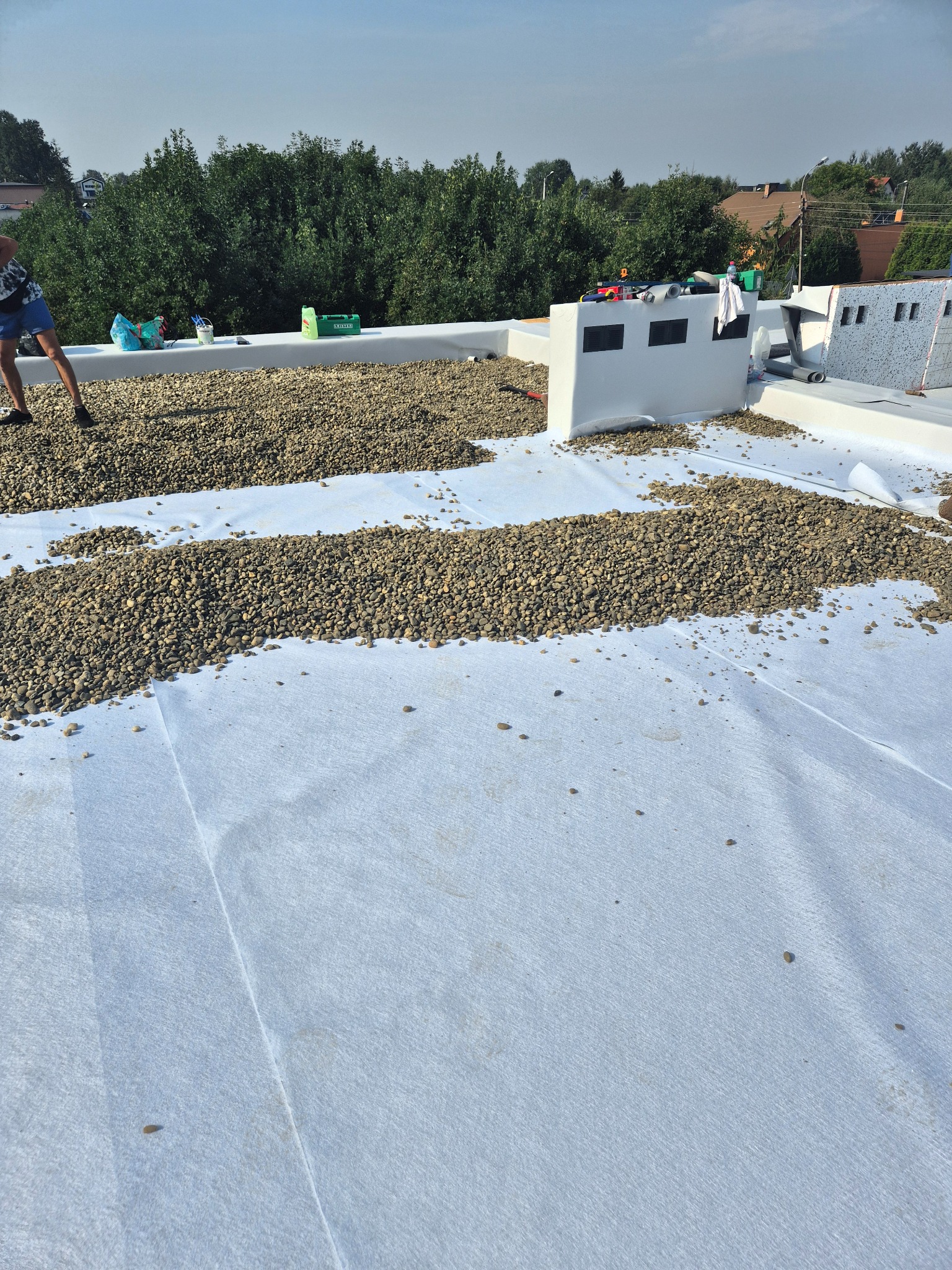Biała membrana dachowa z rozłożonym żwirem na dachu w Zabrzu, widoczne narzędzia i materiały budowlane, drzewa w tle.
