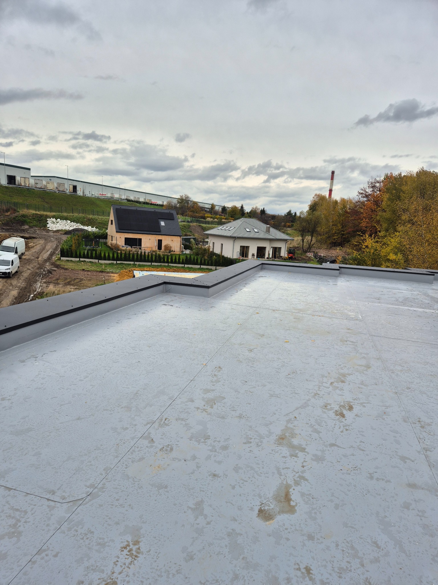 Szary, płaski dach z widocznymi łączeniami, obróbkami blacharskimi i fragmentem krajobrazu z budynkami w tle, sfotografowany w pochmurny dzień.