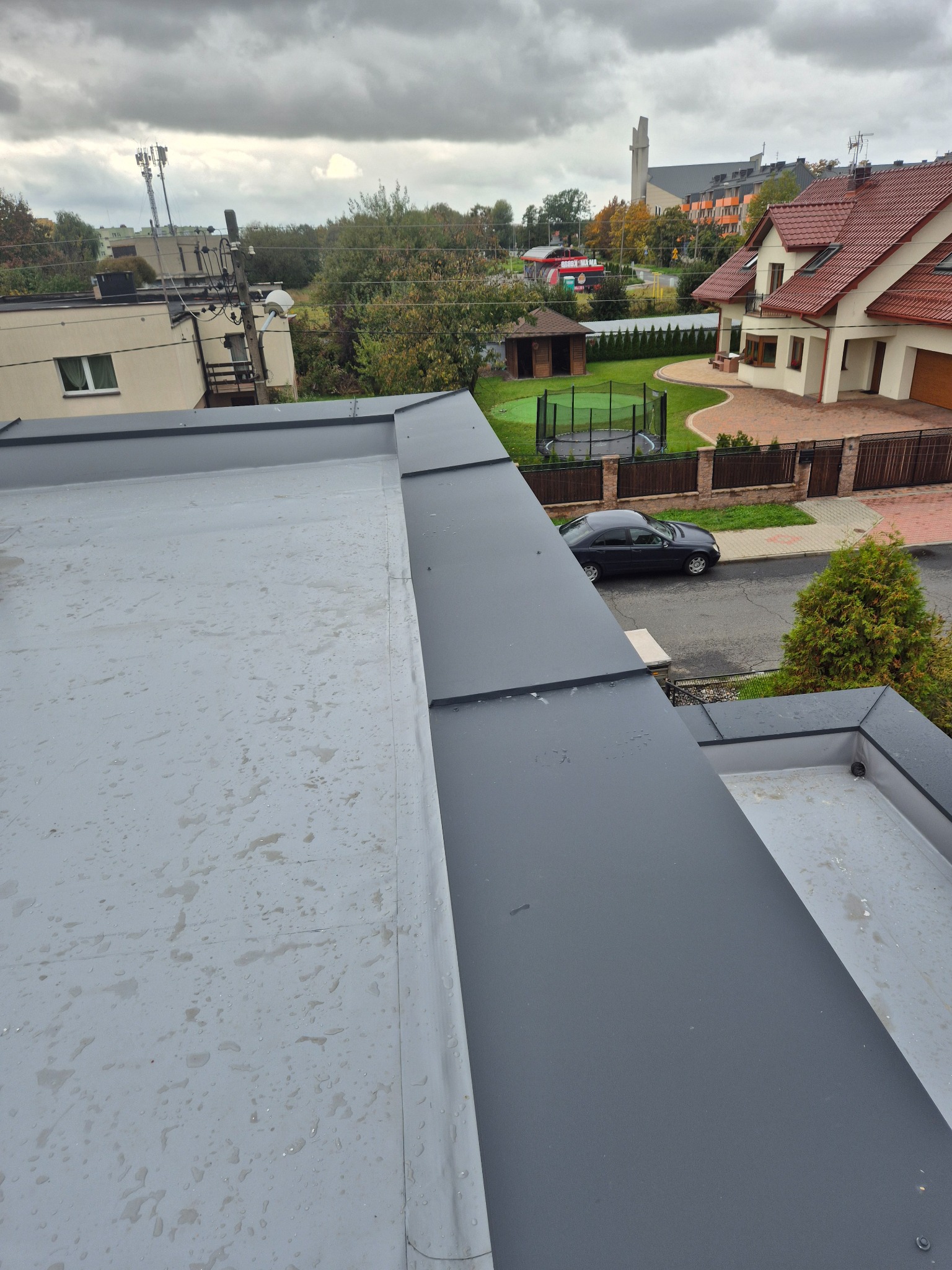 Szary, płaski dach z widocznymi obróbkami blacharskimi w kolorze grafitowym. W tle zabudowa jednorodzinna i kościół. Widok z góry na nową konstrukcję dachu.