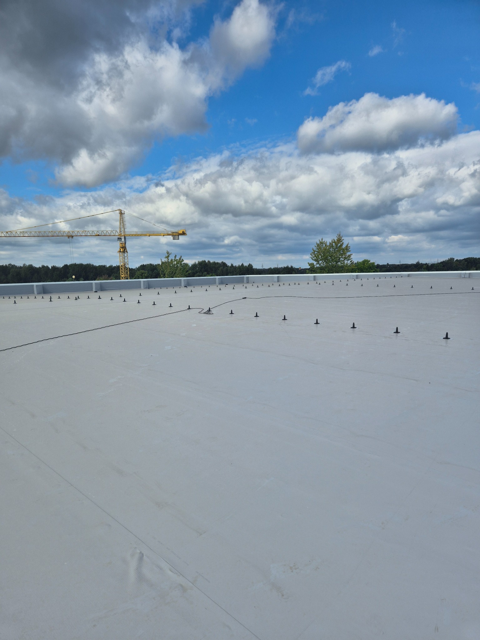 Jasnoszary dach płaski z czarnymi elementami mocującymi, widok z góry. W tle błękitne niebo z chmurami i żółty dźwig budowlany.
