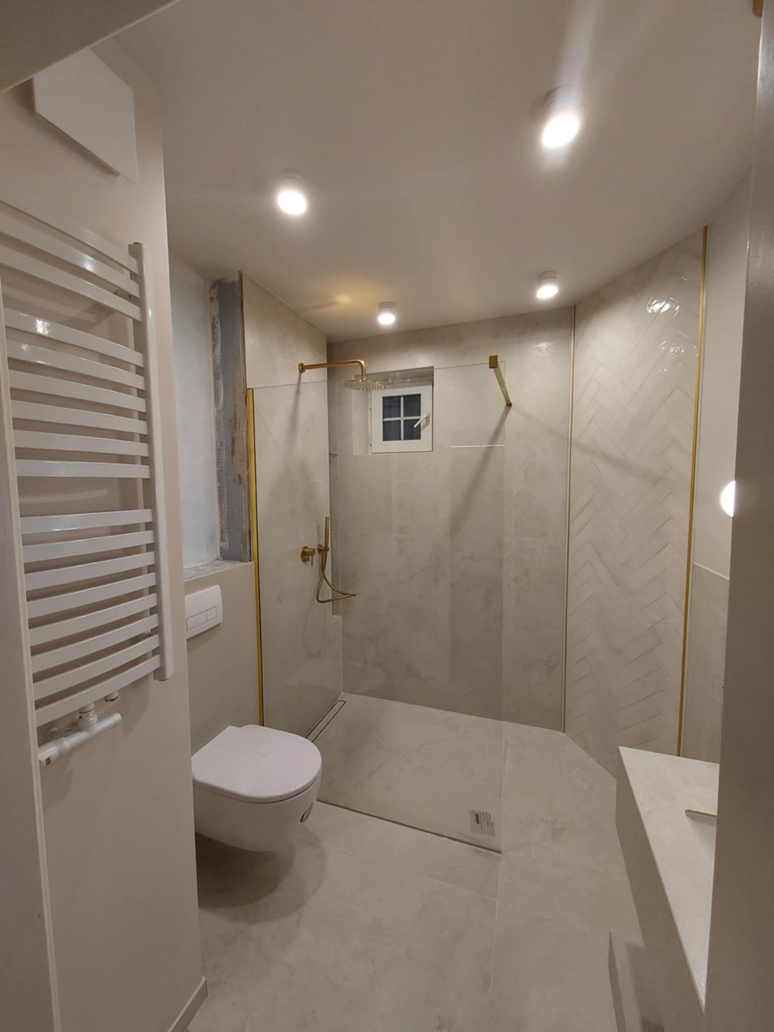 Wykończona łazienka z kabiną prysznicową typu walk-in, beżowymi płytkami w jodełkę i złotymi akcentami. Widoczny grzejnik drabinkowy i toaleta.