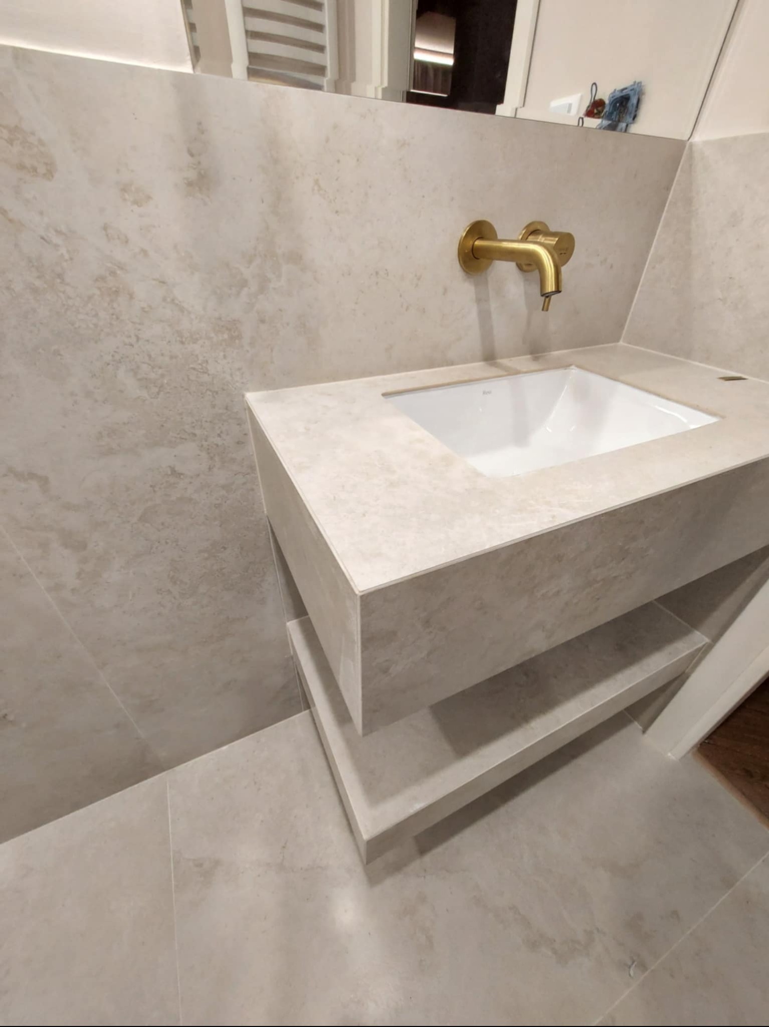 Beżowa łazienka z umywalką nablatową i złotą baterią. Umywalka z półkami, płytki imitujące kamień. Minimalistyczny design, nowoczesne wykończenie.