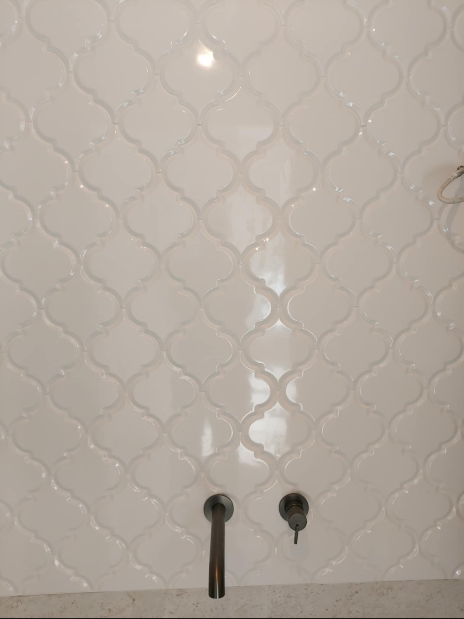 Białe, błyszczące płytki w marokański wzór zdobią ścianę, z widoczną, minimalistyczną armaturą w kolorze grafitowym. Delikatne światło podkreśla fakturę płytek.