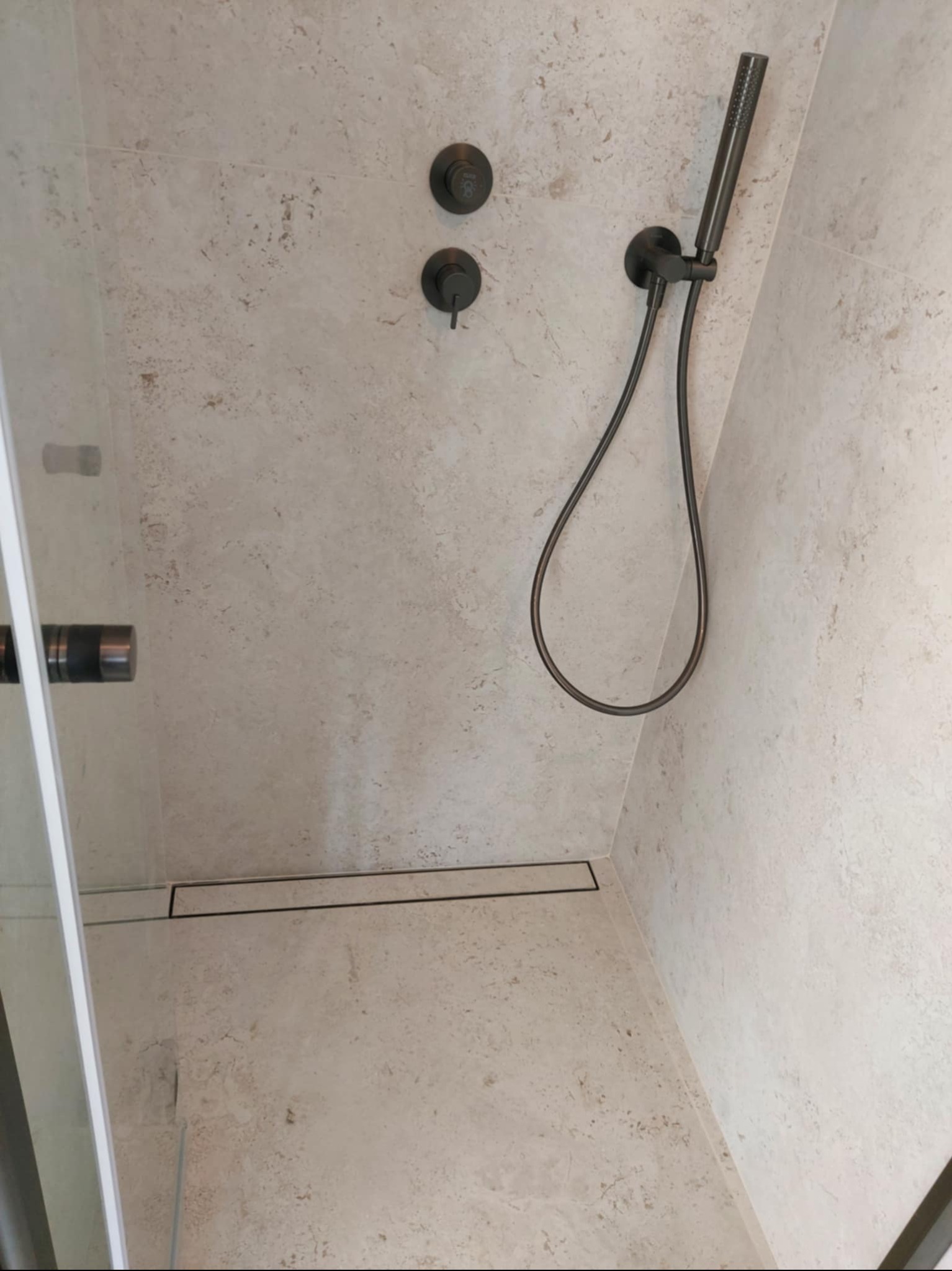 Beżowa kabina prysznicowa z deszczownicą w kolorze grafitowym i odpływem liniowym. Minimalistyczny design, jasne płytki imitujące kamień, szklane drzwi.