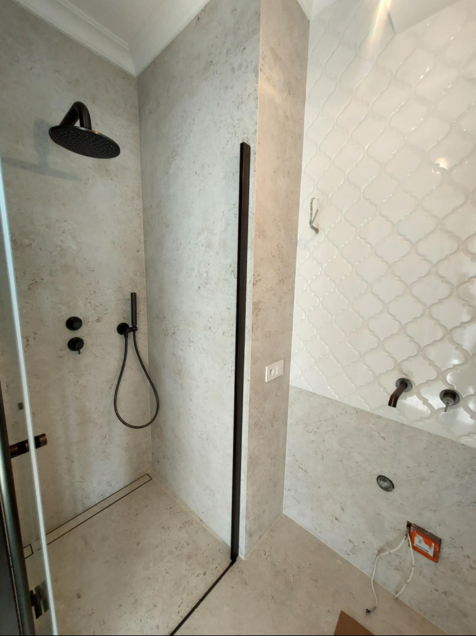 Nowoczesna kabina prysznicowa z czarną armaturą i odpływem liniowym, wykończona płytkami w odcieniach szarości i bieli. Widoczny fragment ściany z dekoracyjną mozaiką.