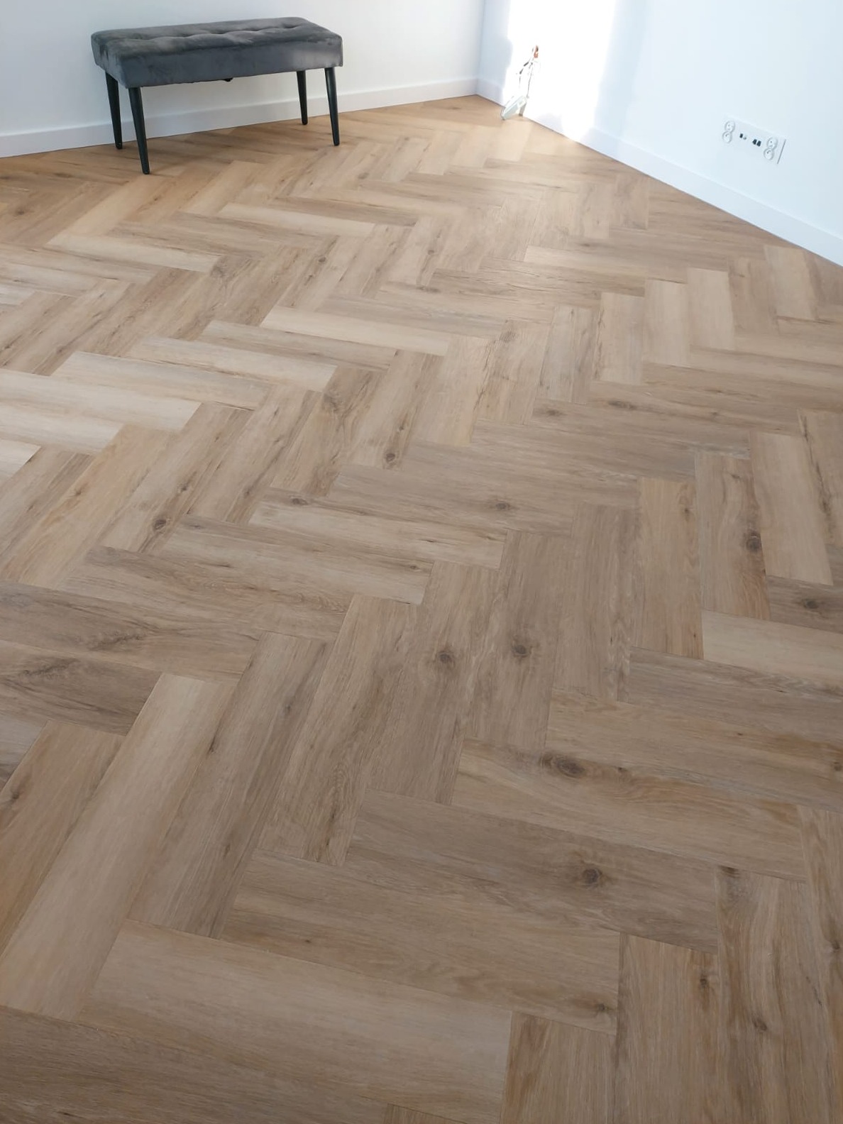 Jasna, drewnopodobna podłoga ułożona w jodełkę, z szarą pufą w rogu pokoju. Widoczne gniazdka elektryczne na białej ścianie i fragment okna z wpadającym światłem.