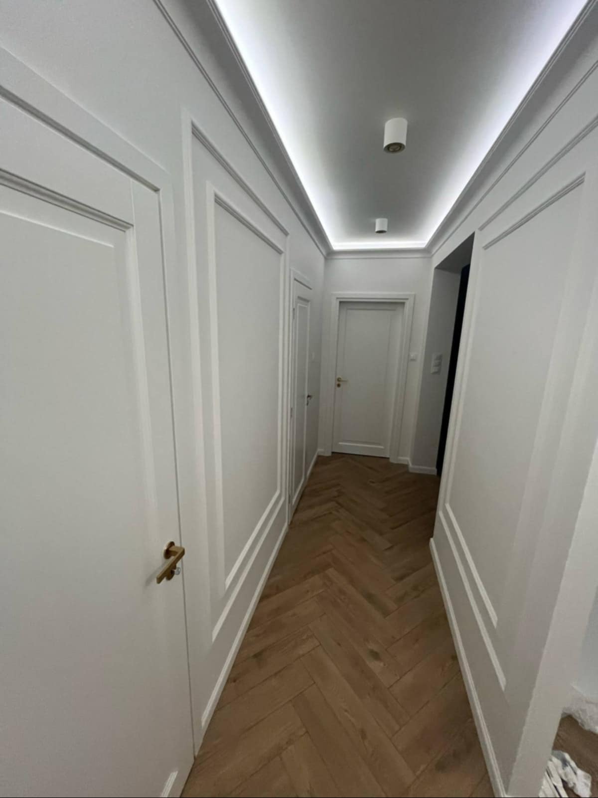 Biały korytarz z parkietem w jodełkę, ozdobnymi listwami ściennymi i subtelnym oświetleniem sufitowym. Widok na drzwi i wnętrze.