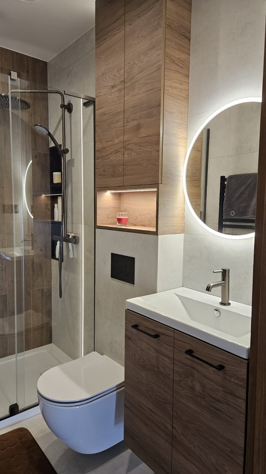 Nowoczesna łazienka z kabiną prysznicową, wiszącą toaletą i szafką z umywalką w odcieniach brązu i szarości. Okrągłe lustro z podświetleniem LED.