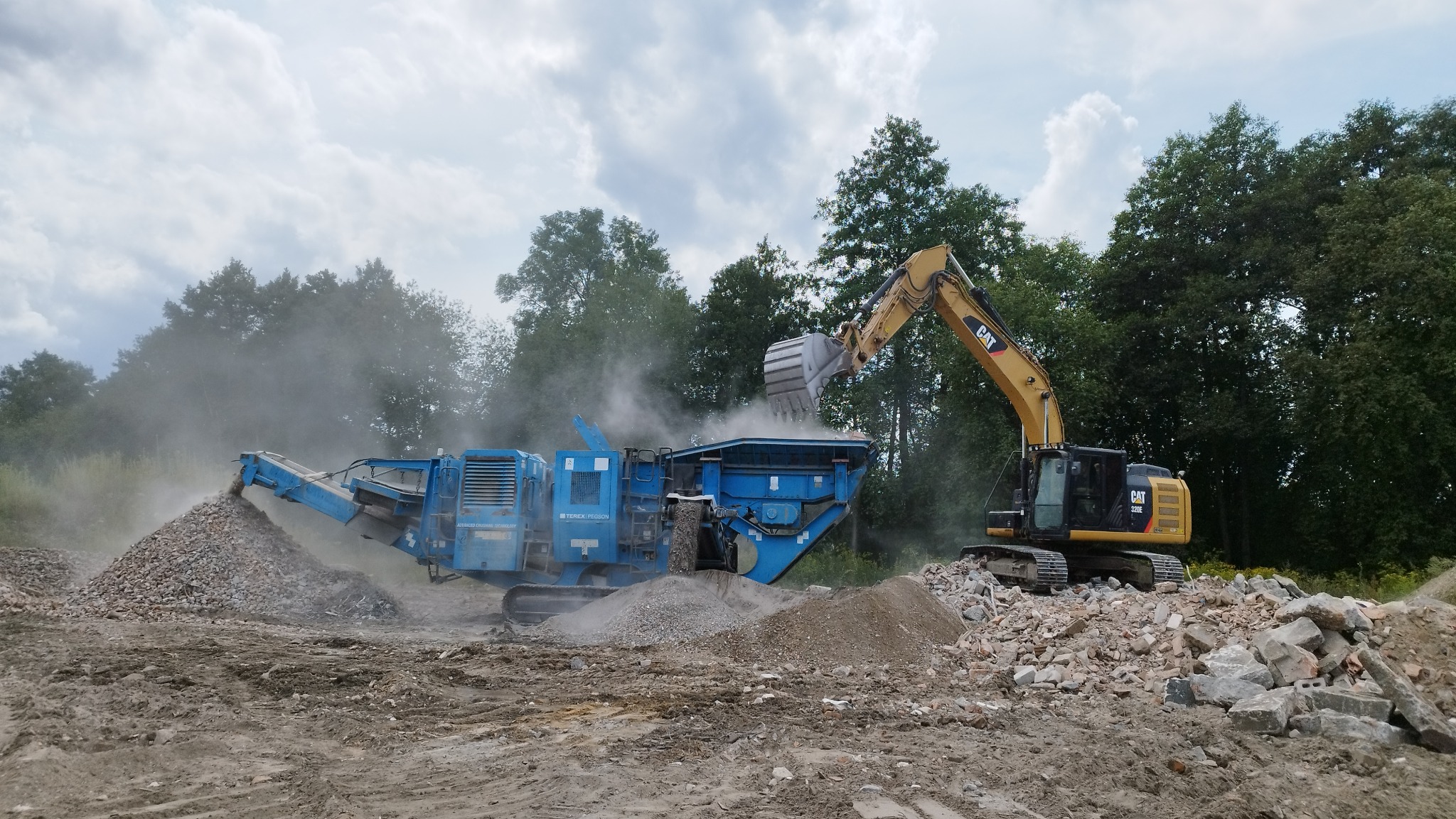 Żółta koparka CAT 320E podnosi gruz i wrzuca do niebieskiego kruszarki Terex Pegson na tle drzew i pochmurnego nieba.