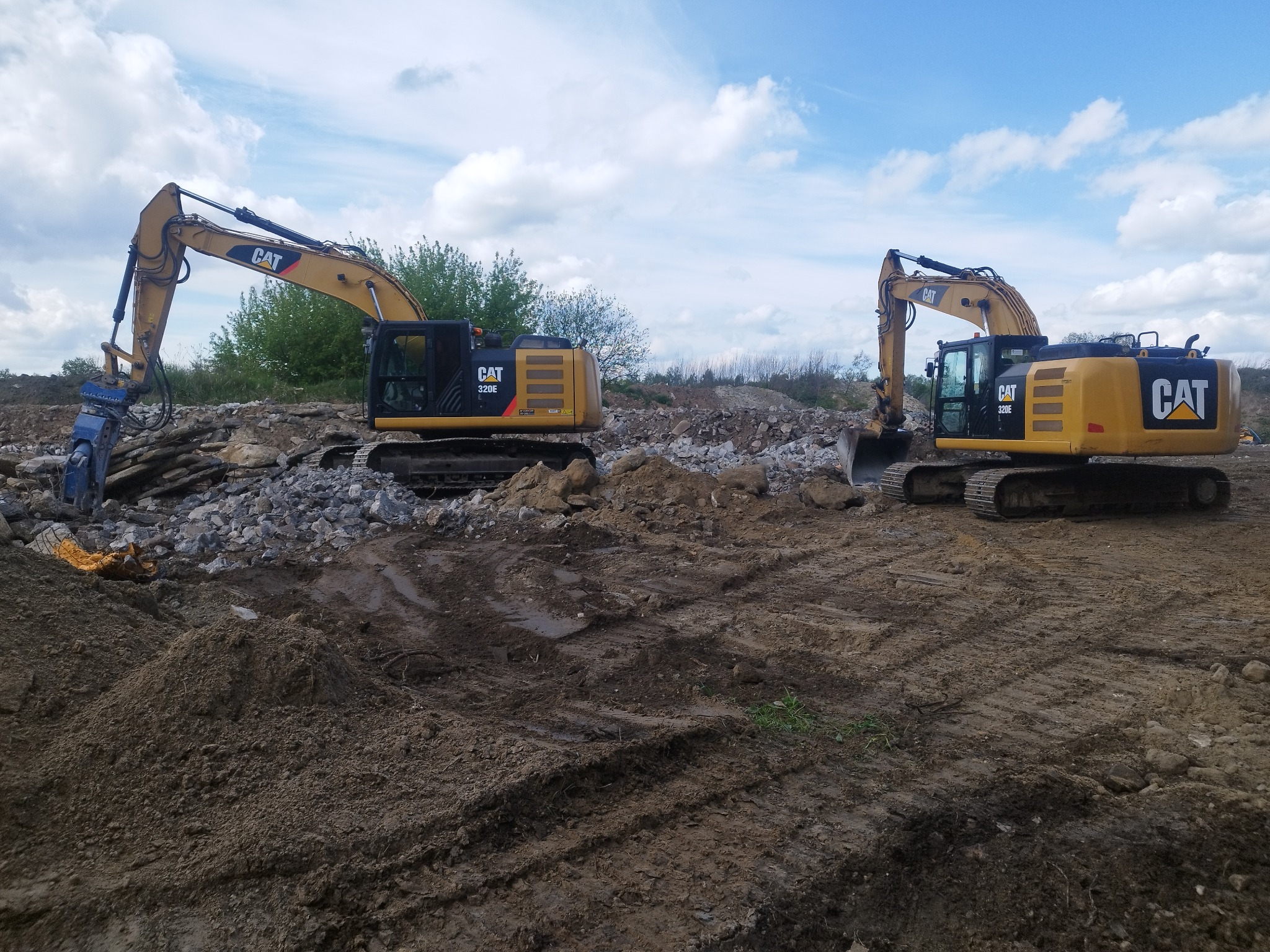 Dwa żółte, gąsienicowe koparki CAT 320E na placu budowy, jedna z młotem hydraulicznym kruszy gruz, druga z łyżką stoi na ziemi, w tle zielona roślinność i błękitne niebo z chmurami.