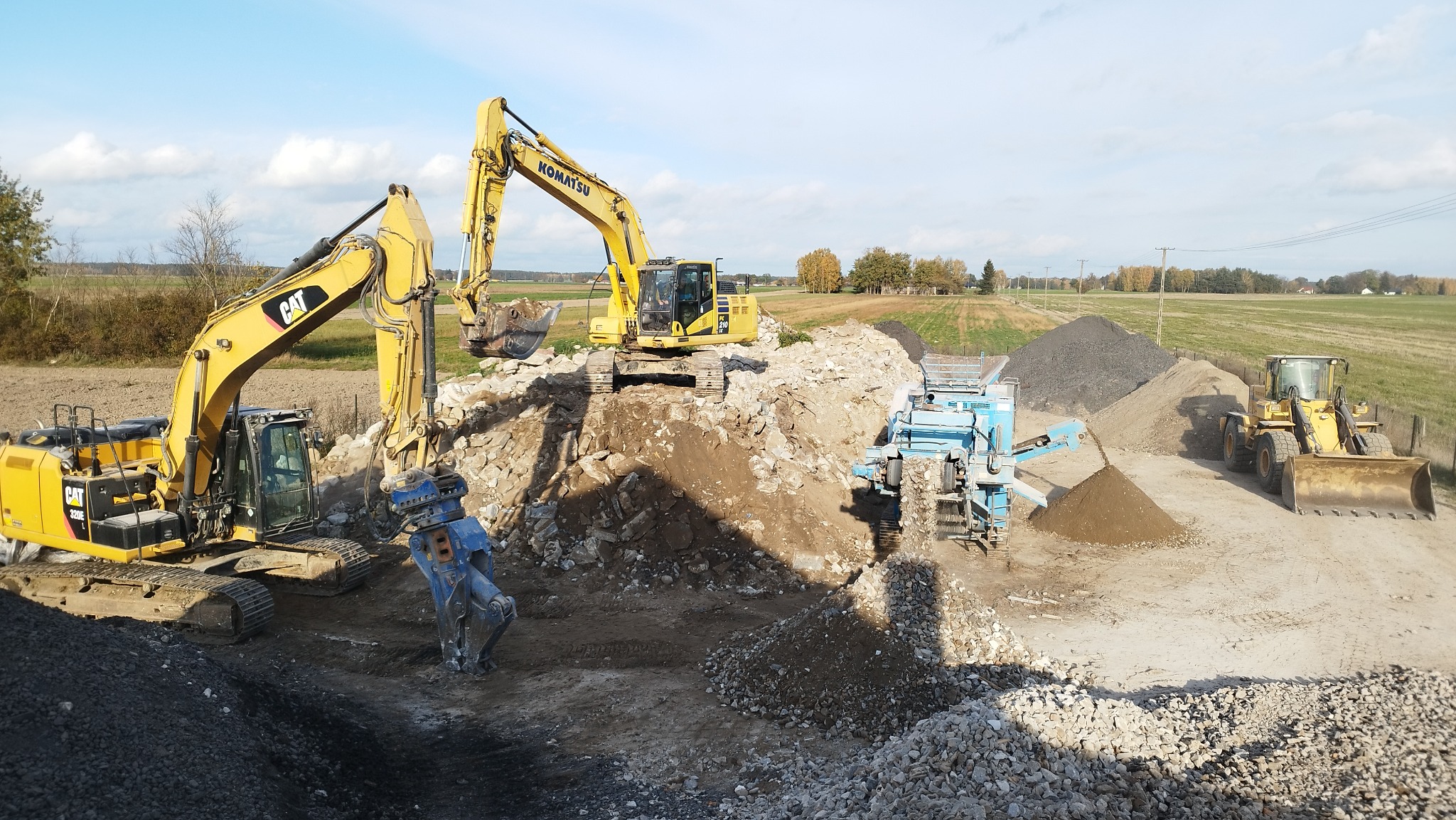 Dwa żółte koparki CAT i Komatsu pracują na placu budowy, jedna z niebieskim młotem hydraulicznym, w tle hałdy kruszywa i przesiewacz, w tle widoczne pola i drzewa pod błękitnym niebem.