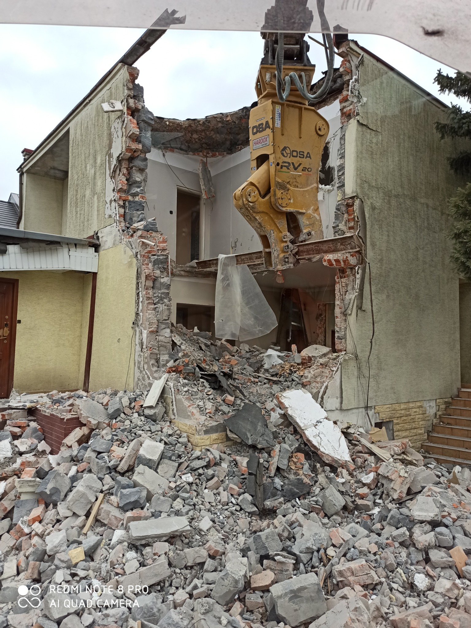 Żółty sprzęt budowlany OSA RV20 kruszy fragment ściany domu, na ziemi leżą gruzy i cegły, widoczne zniszczone wnętrze budynku.