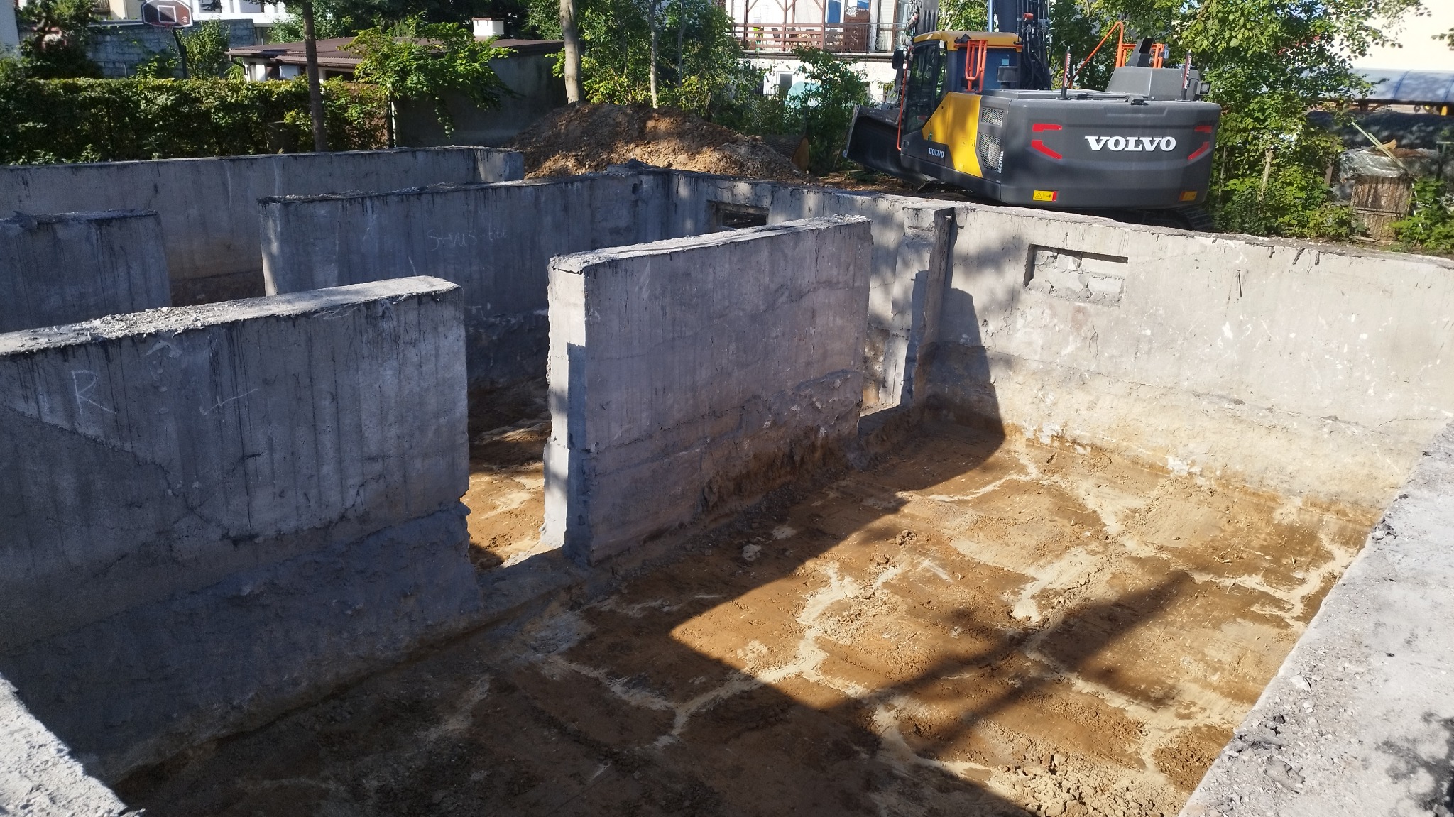 Zalane betonowe ściany fundamentowe z wykopem w środku, na zewnątrz ziemia i koparka Volvo. Widoczne ślady po zalewaniu betonu.