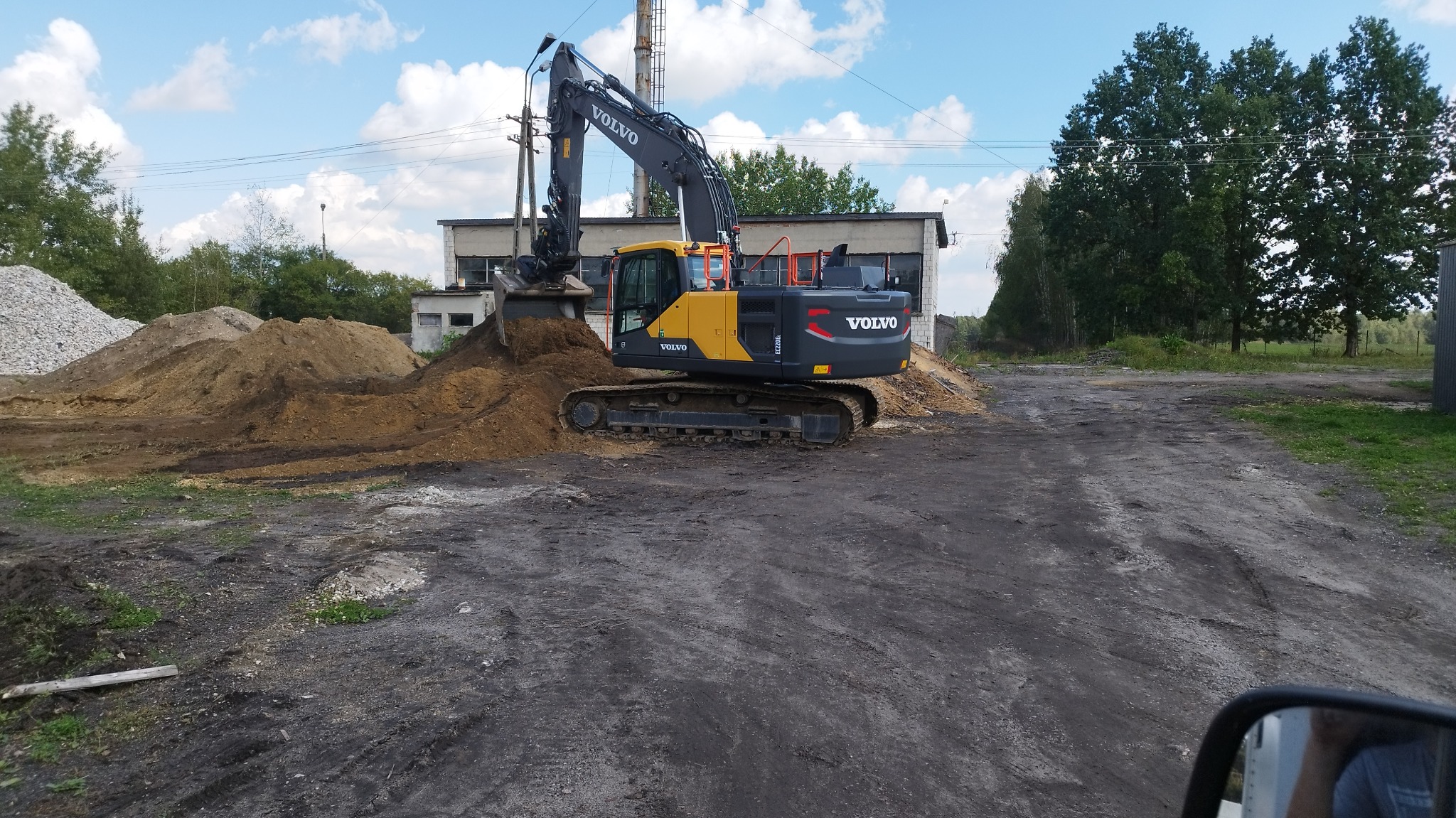 Żółto-szara koparka Volvo na placu budowy, z hałdami ziemi i gruzu w tle. Widoczne budynki przemysłowe i błękitne niebo z chmurami. Praca w toku.