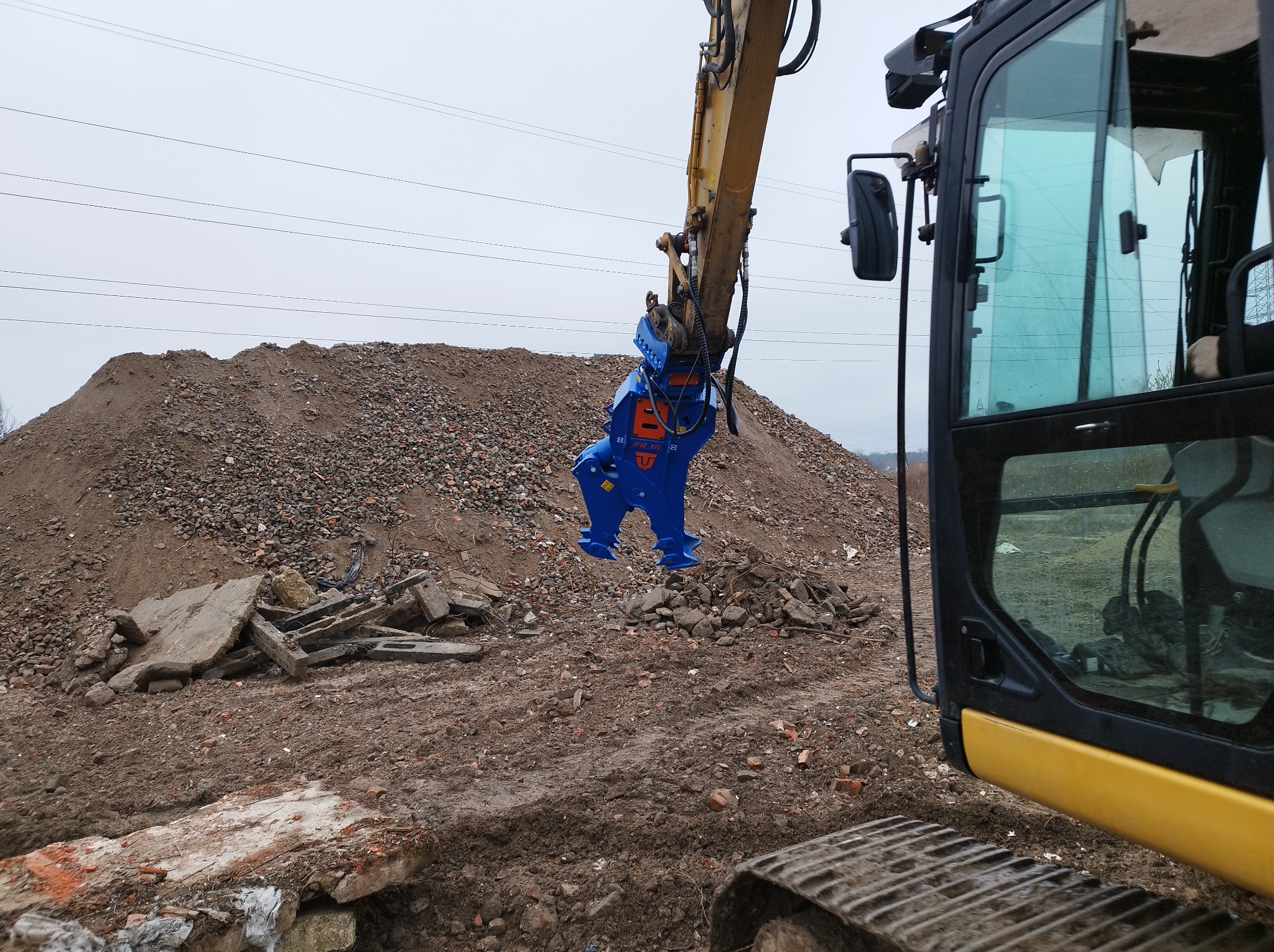 Żółta koparka z niebieskim osprzętem do kruszenia betonu na tle sterty gruzu i ziemi. Widok z boku kabiny, pochmurne niebo.