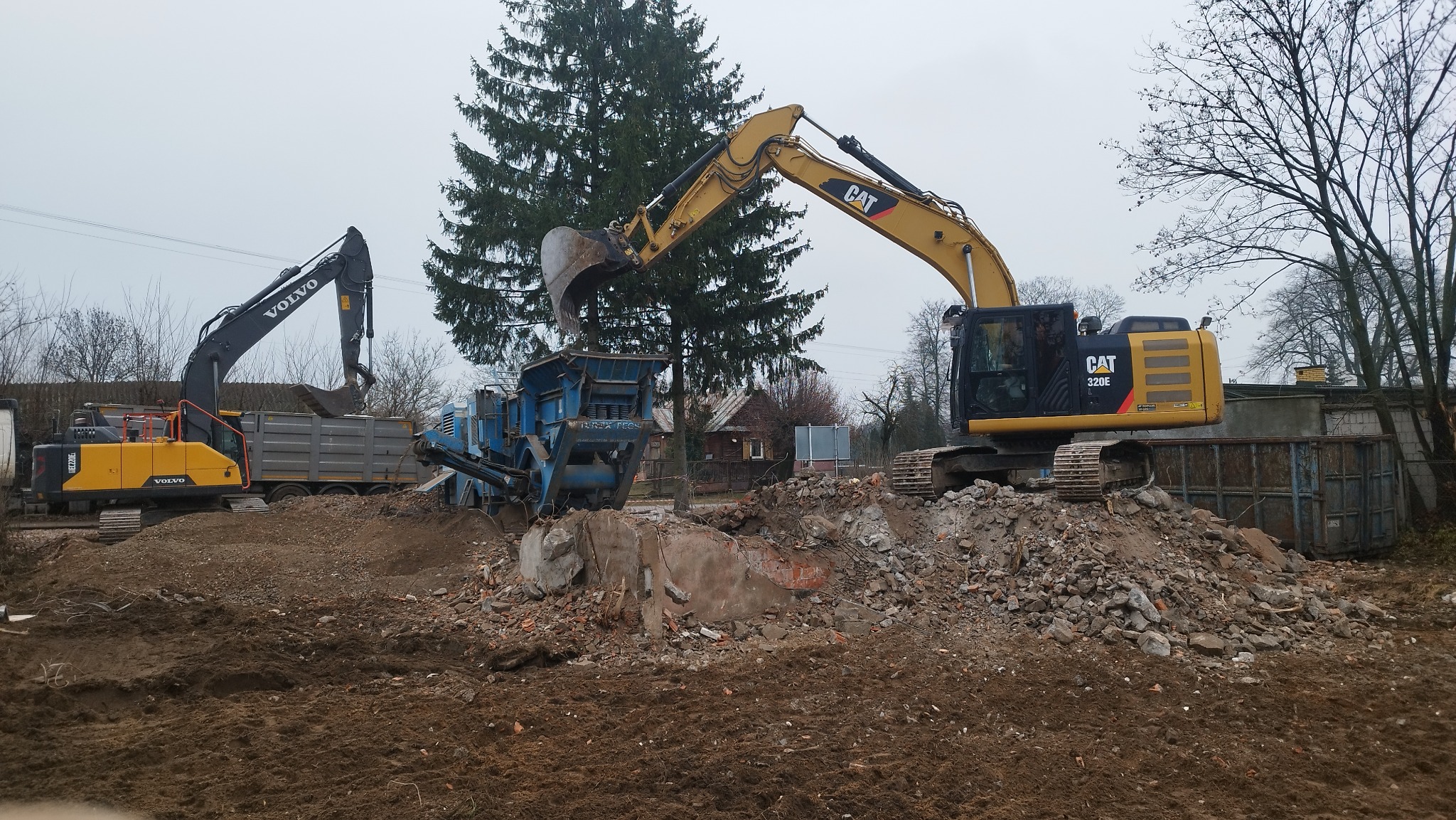 Żółty CAT 320E i czarno-żółty Volvo kopią gruz z rozbiórki do niebieskiego rozdrabniacza, w tle drzewa i budynki. Praca wre na placu budowy.