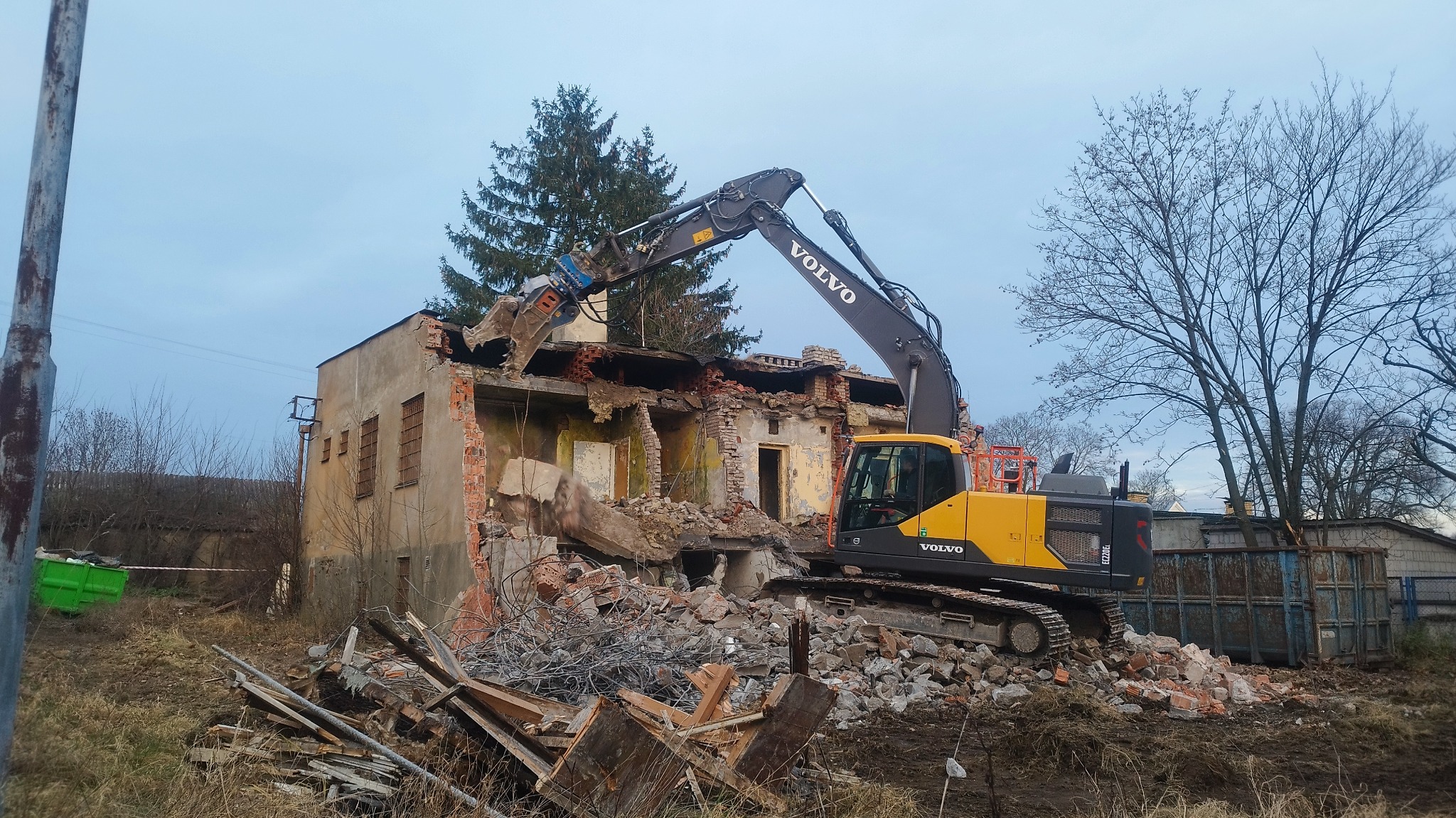Żółta koparka Volvo na gąsienicach rozbiera budynek z cegły. Widoczne gruzy i fragmenty ścian. Praca w toku, pochmurne niebo w tle.