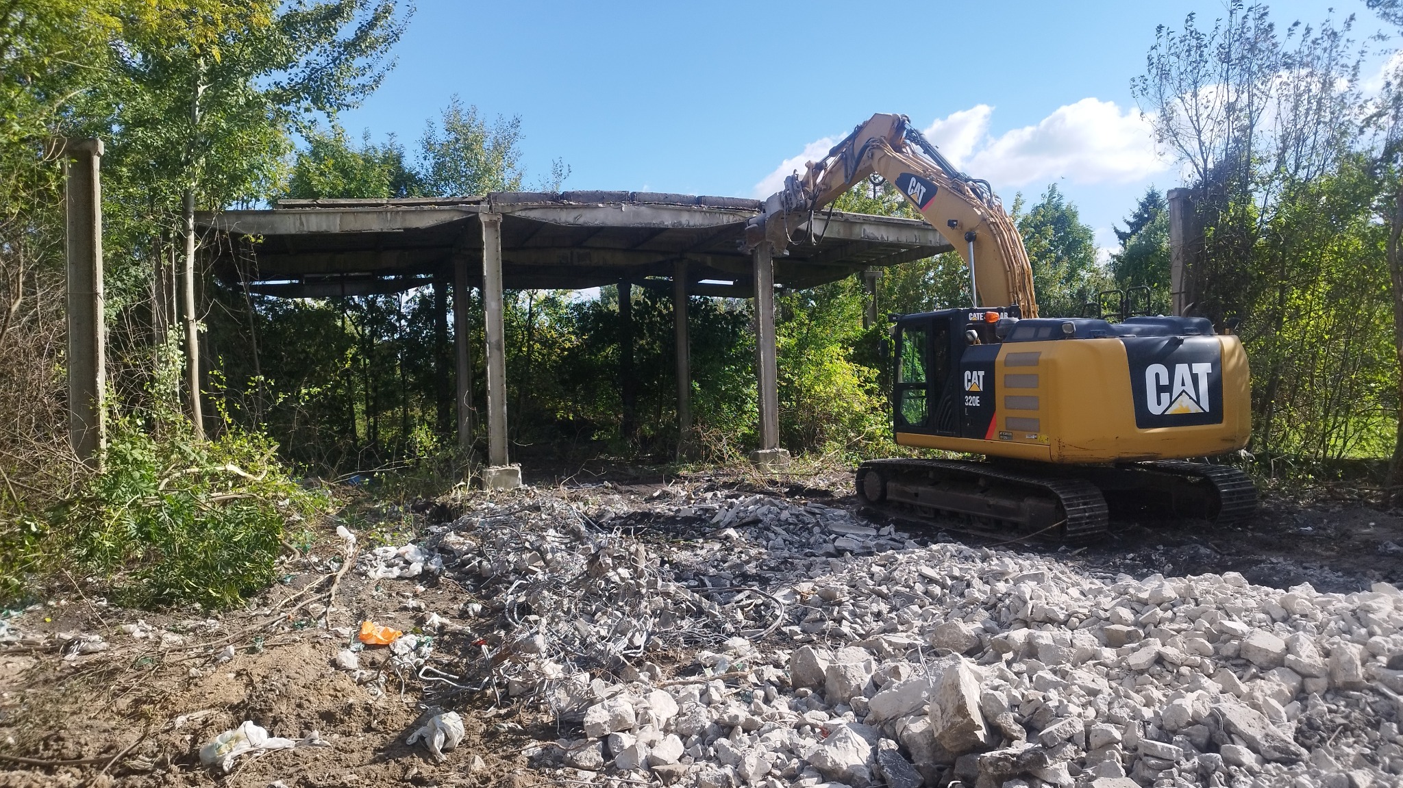 Żółta koparka CAT 320E w trakcie rozbiórki betonowej konstrukcji z zarośniętymi filarami, gruzowisko na pierwszym planie, błękitne niebo w tle.