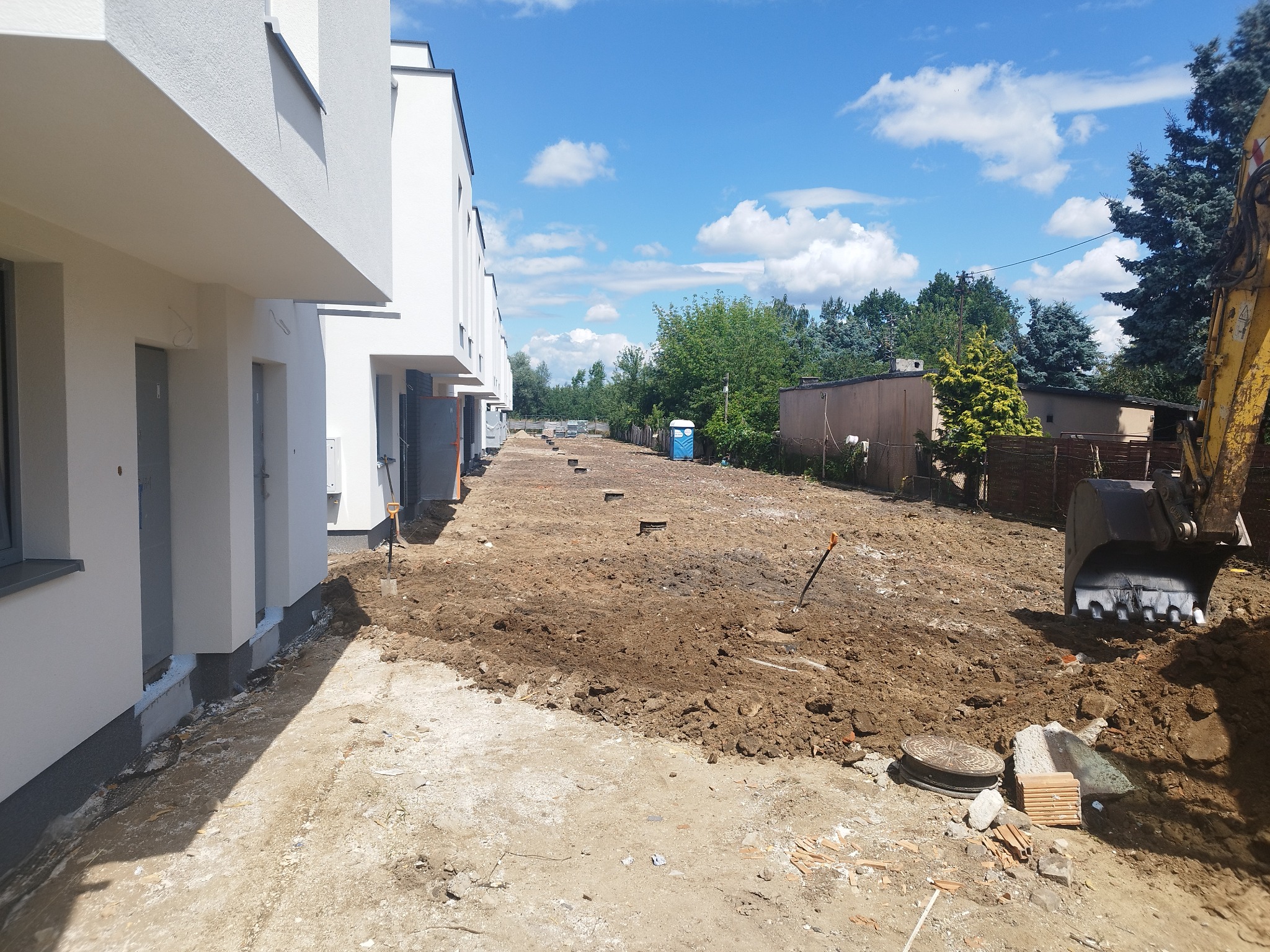 Żółta koparka na placu budowy, przygotowany teren pod budowę szeregu domów jednorodzinnych, widoczne studzienki kanalizacyjne, przenośna toaleta i łopata wbita w ziemię.